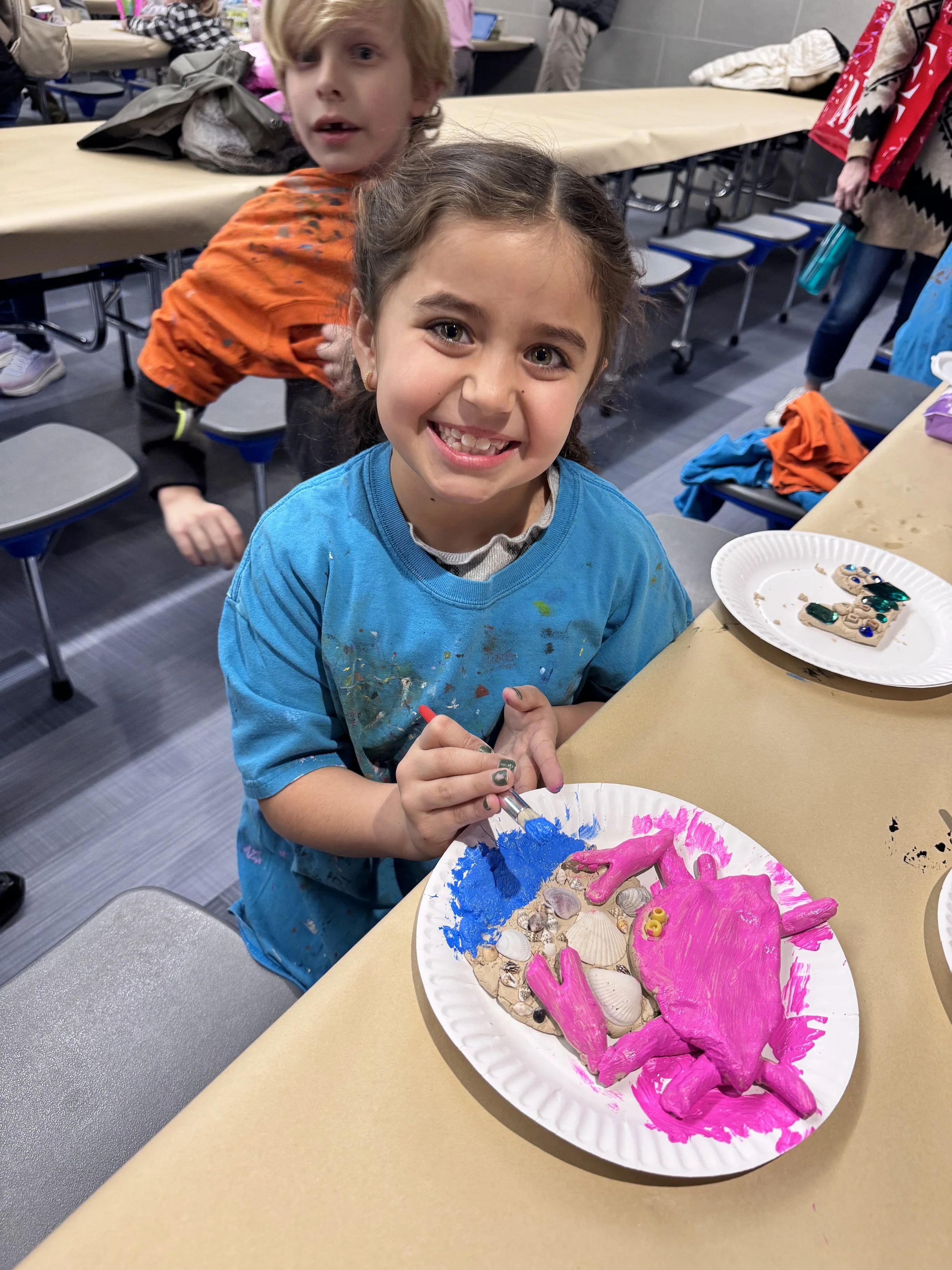 A young girl smiling at the camera while painting a pink dinosaur on a shell collage with blue paint, sitting at a table covered with arts and crafts supplies, with a boy in an orange shirt and other children in the background.