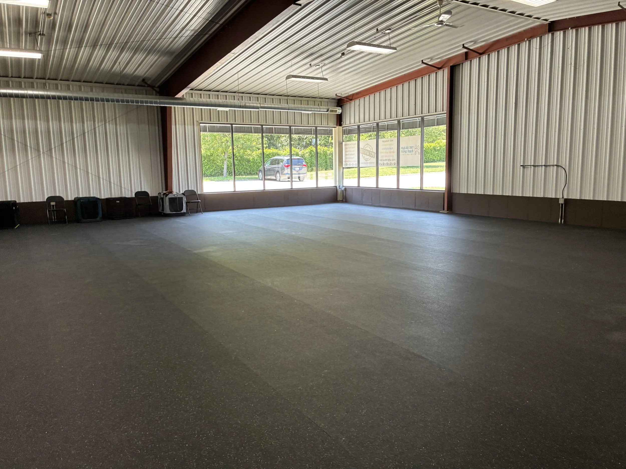 Empty indoor room with carpeted floor, metal walls, and large windows facing outside. There are a few chairs and equipment against the wall.