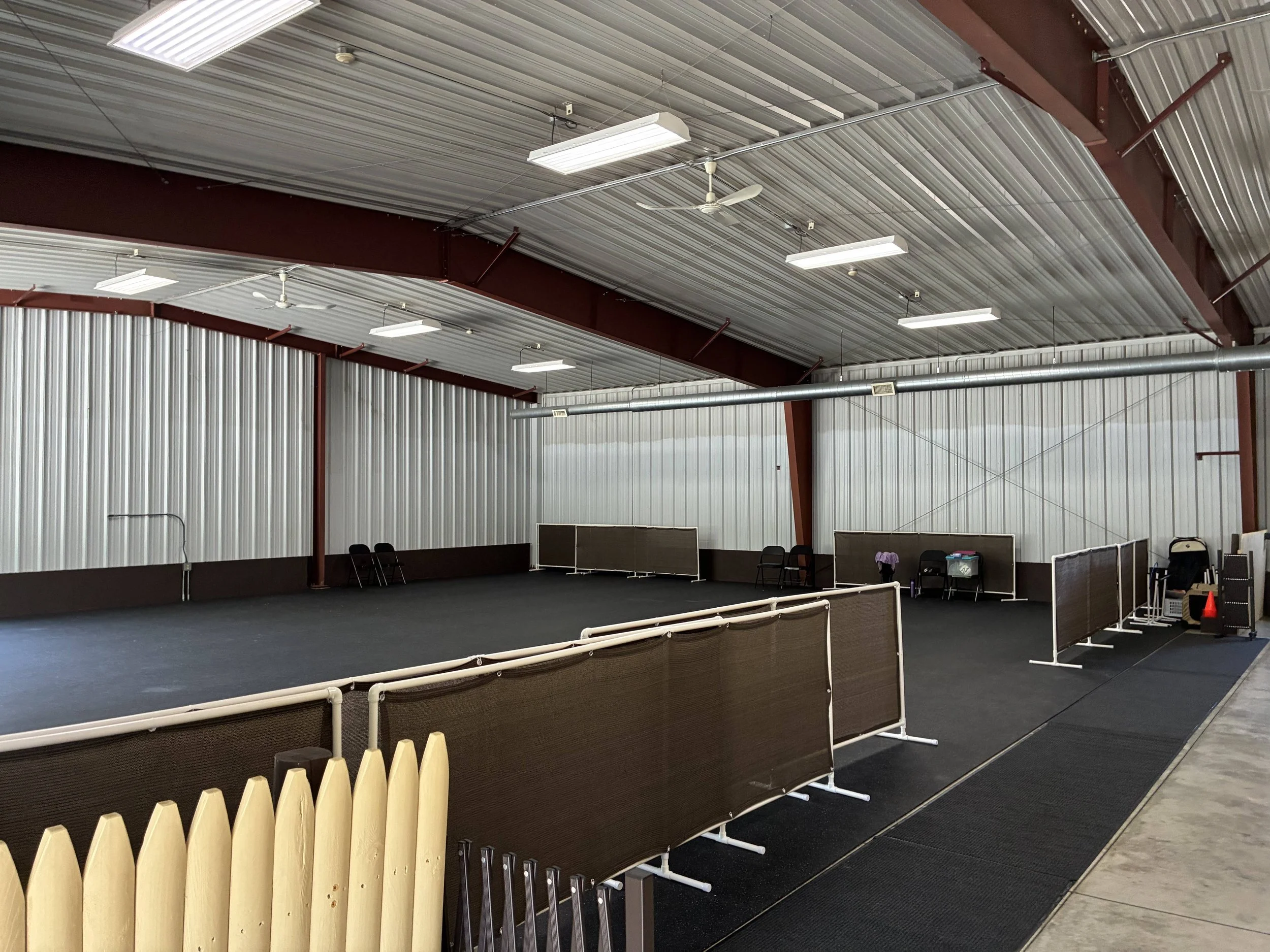 Indoor sports facility with a black turf playing field, beige wooden fencing, and overhead fluorescent lighting, ceiling fan, and metal beams.