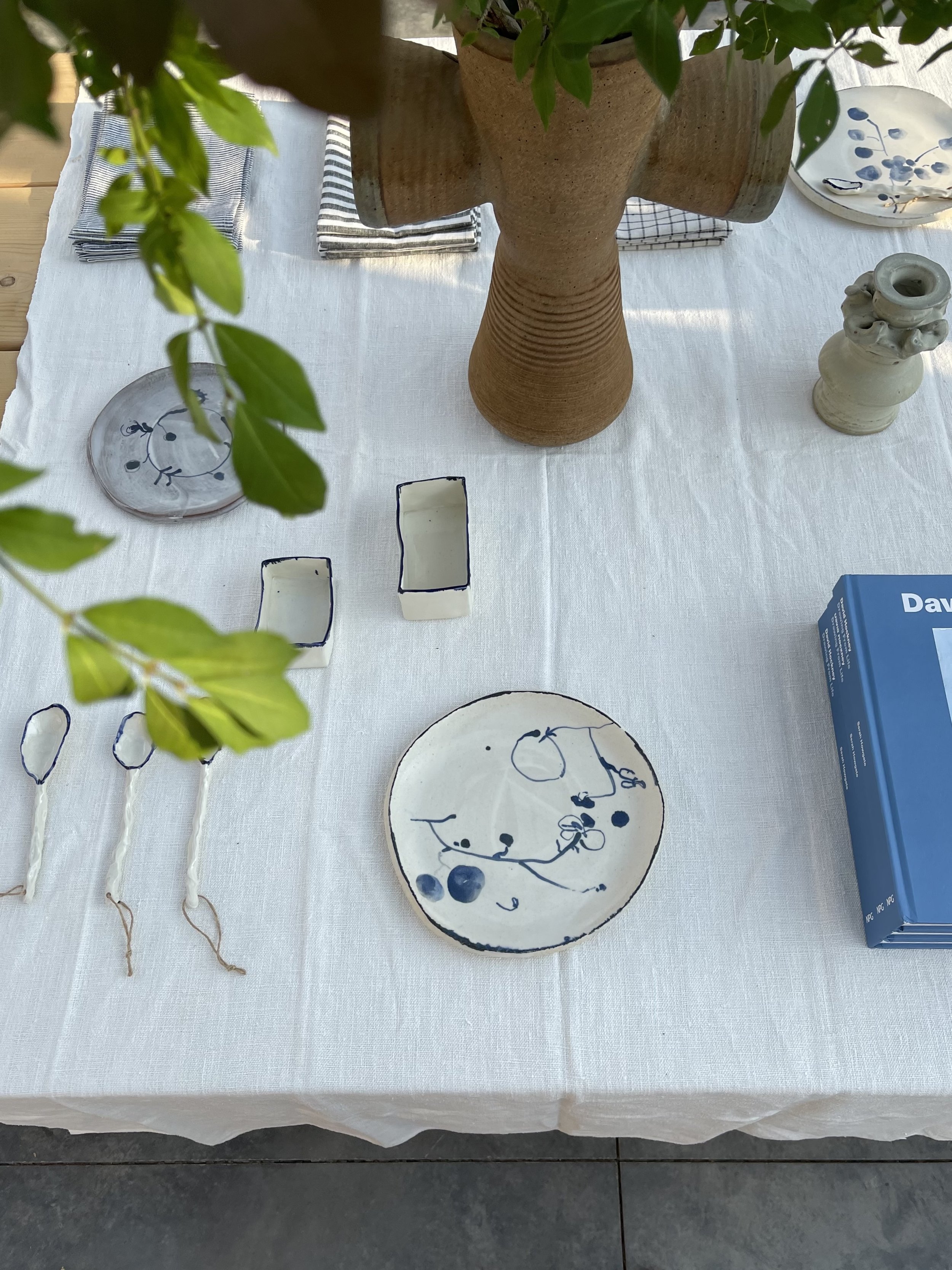 A table with a white tablecloth displaying ceramic plates and bowls with blue abstract designs, small candle holders, a brown ceramic vase, and folded checkered cloths, with some foliage in the foreground.