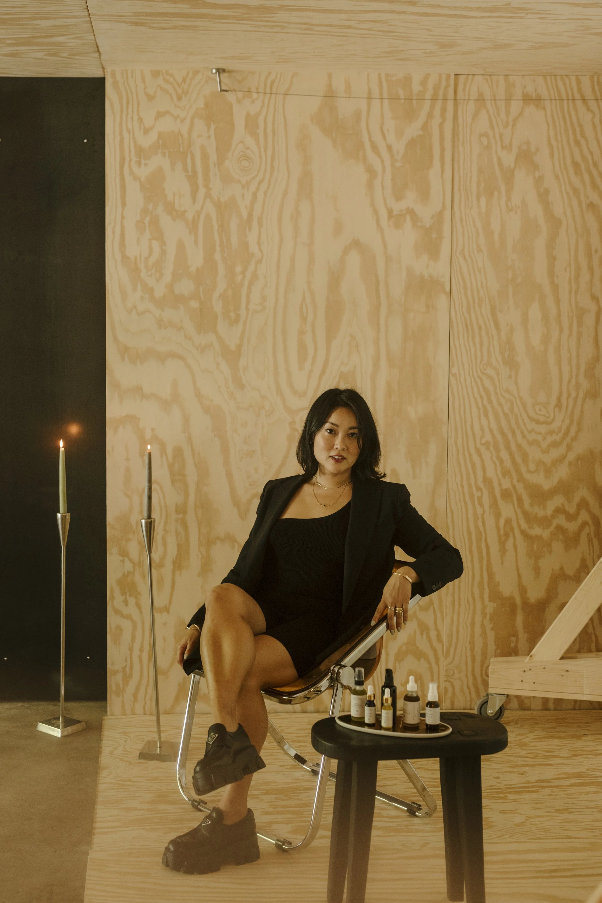 Woman sitting on a chair in a modern, minimalistic room with wood-paneled walls, surrounded by skincare or beauty product bottles on a small table.