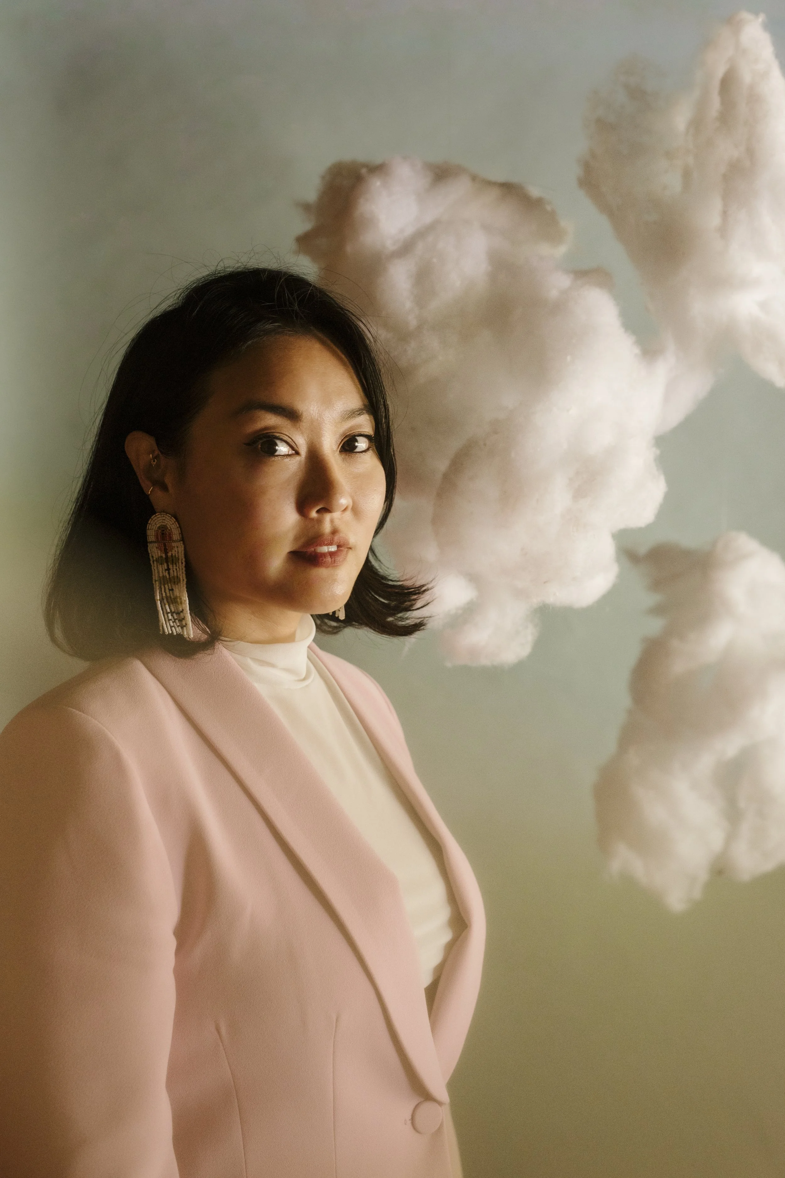 A woman with black hair and earrings, wearing a light pink blazer over a white turtleneck, standing in front of a background with cotton candy clouds.