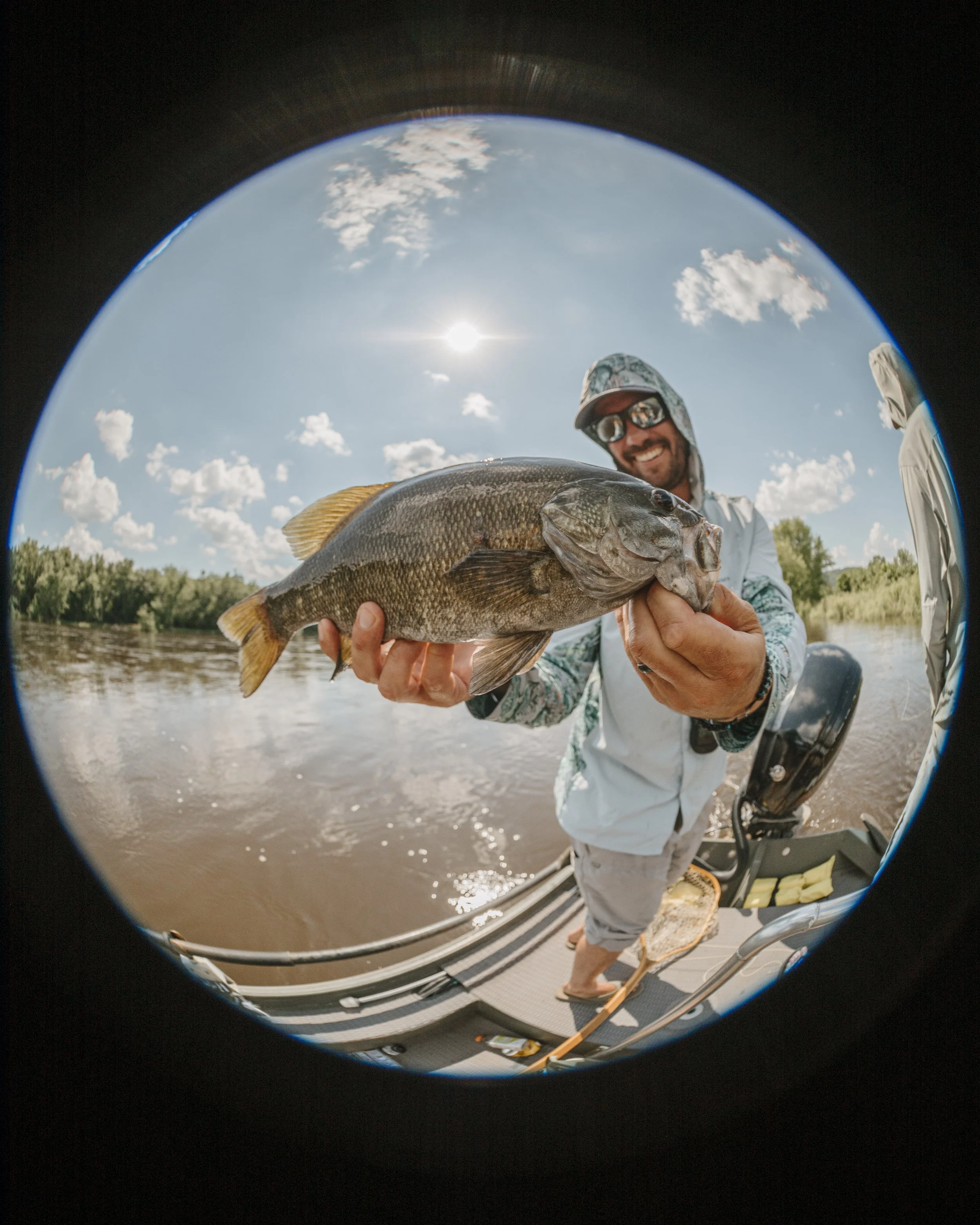 Man holding a large fish on a boat in a river, with a bright sky and scattered clouds.