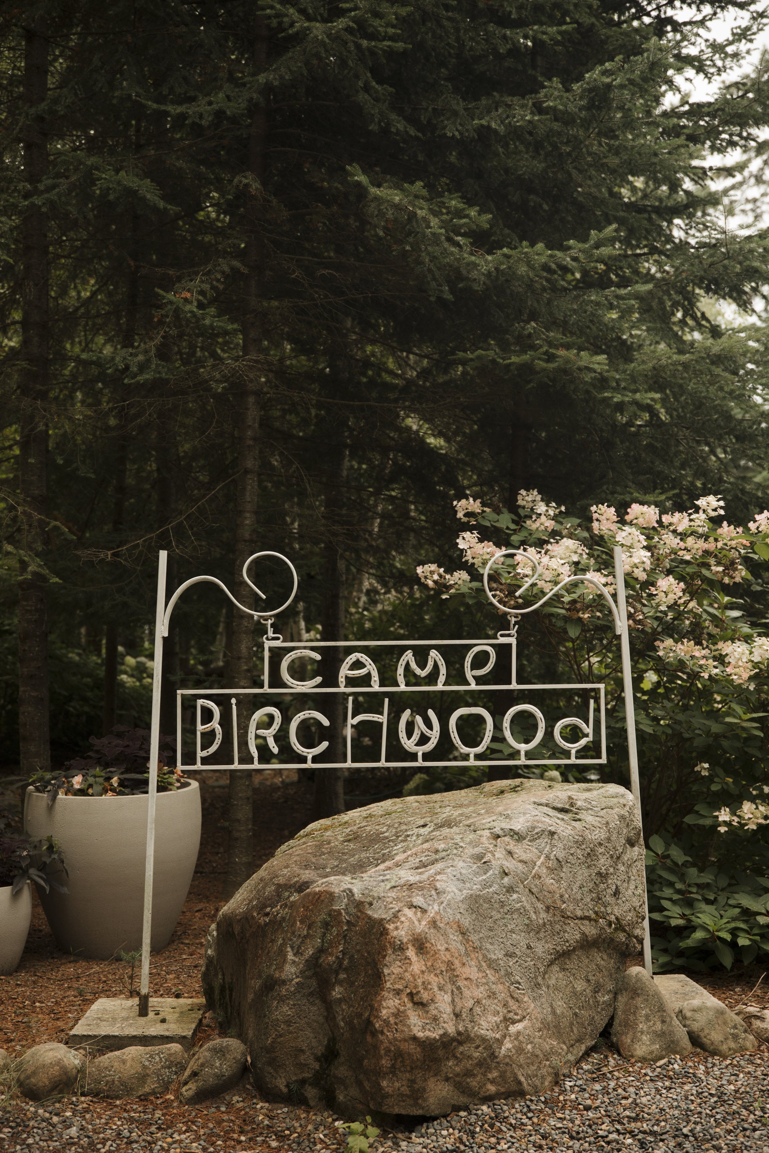 A white metal sign on a large rock reads 'Camp Birchwood,' surrounded by potted plants and flowering bushes in a wooded area.