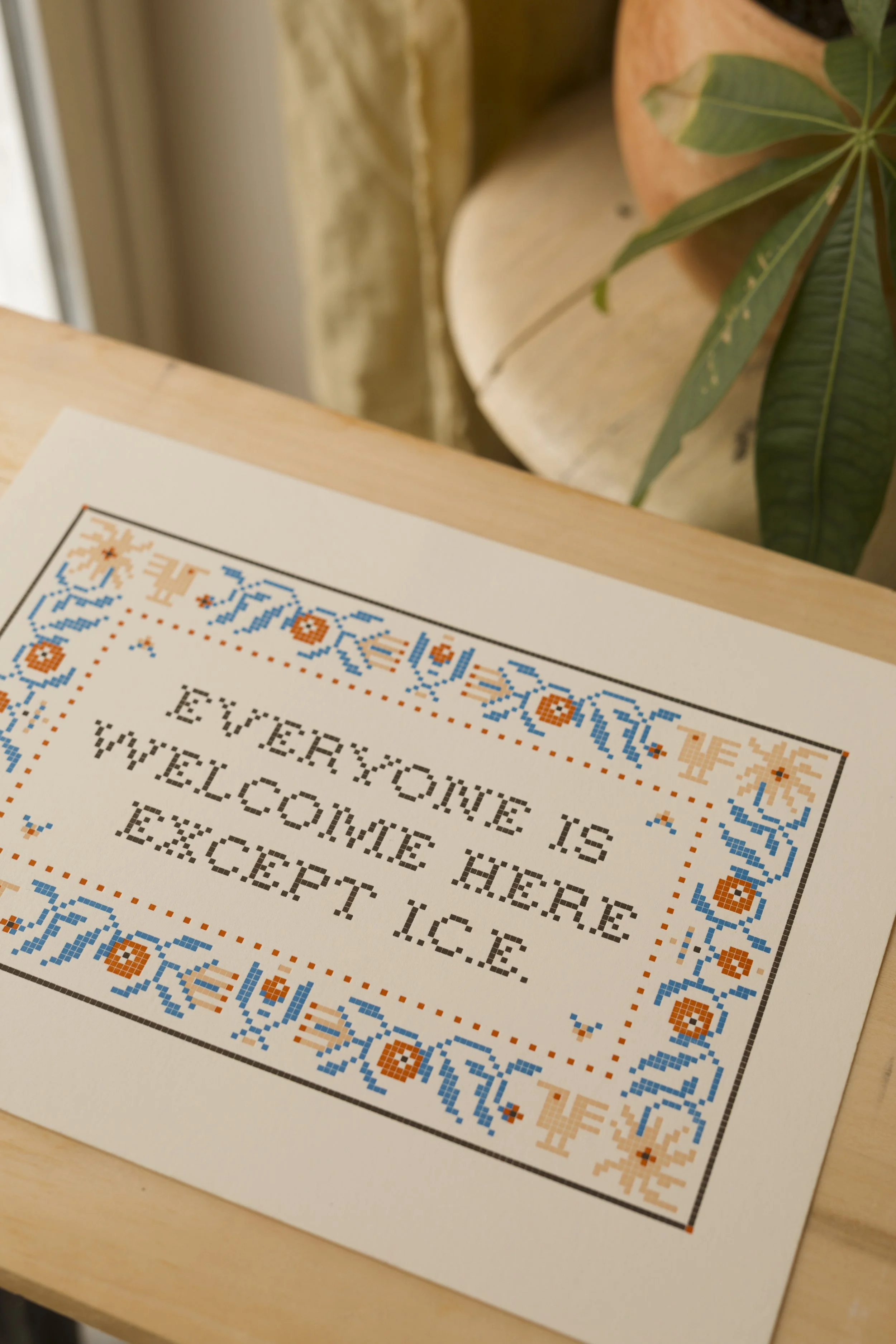 Cross-stitch sampler with the quote 'Everyone is welcome here, welcome hug, respect & love.' decorated with a floral border in blue, orange, and beige. Part of a potted plant is visible in the background.