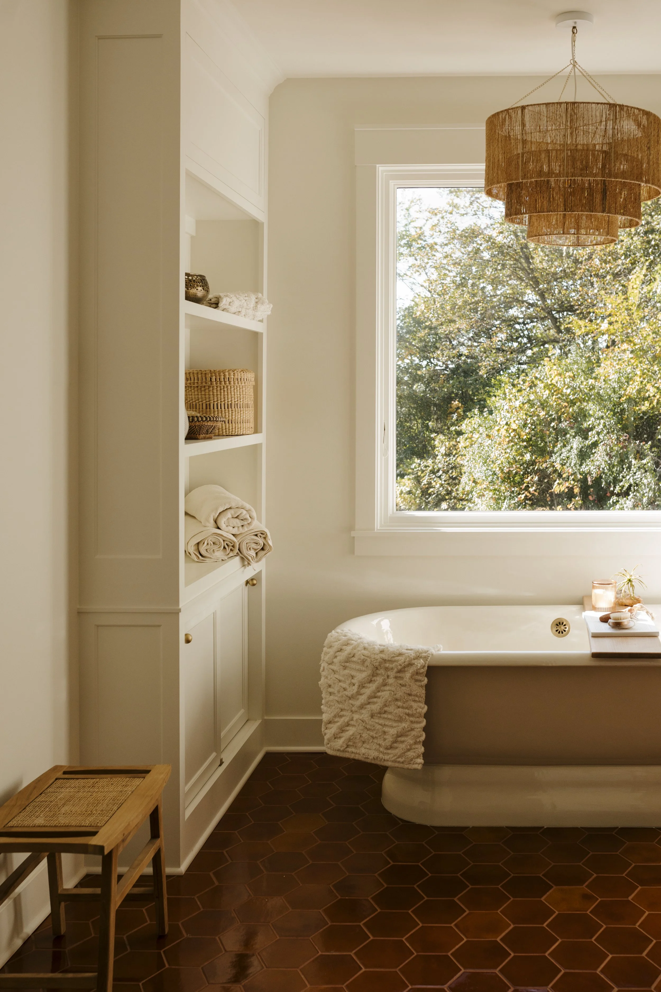 Bright bathroom with a large window showing trees outside, a white bathtub with a textured towel draped over the side, a built-in white shelf with folded towels and baskets, a wooden stool, and a layered brown pendant light hanging from the ceiling.