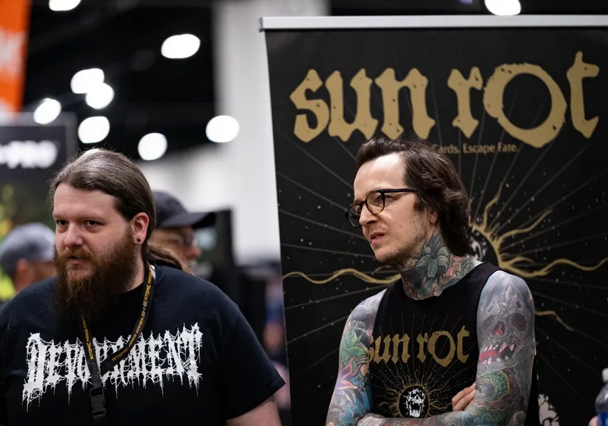 Two men standing at a convention, one with a beard wearing a black t-shirt with white text, and the other with tattoos on his arms and neck wearing glasses and a black sleeveless shirt with a design. Behind them is a black banner with gold text that 