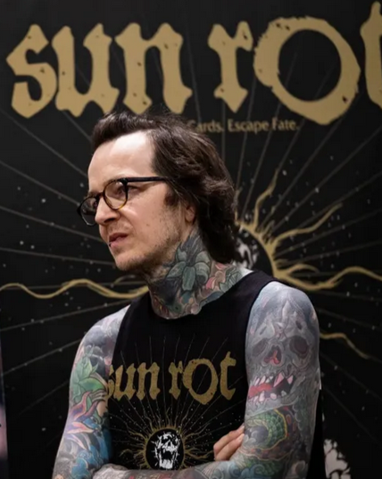 A man with glasses and tattoos on his arms and neck standing in front of a black backdrop with the Allah Ot logo and skull design.
