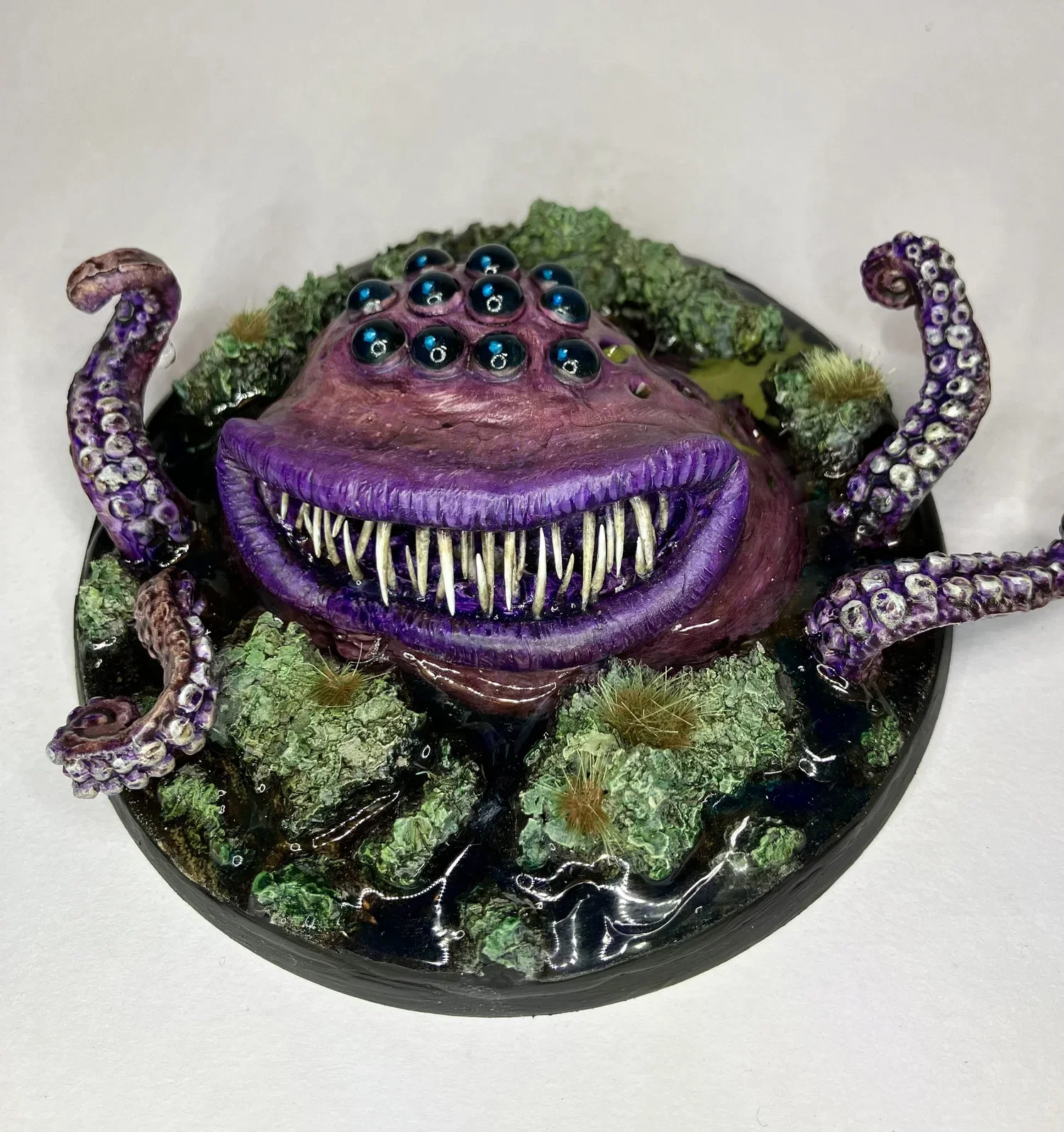 A detailed, colorful sculpture of a fantastical creature resembling a tentacled monster with multiple eyes and sharp teeth, set on a decorative base with green moss and rocks.