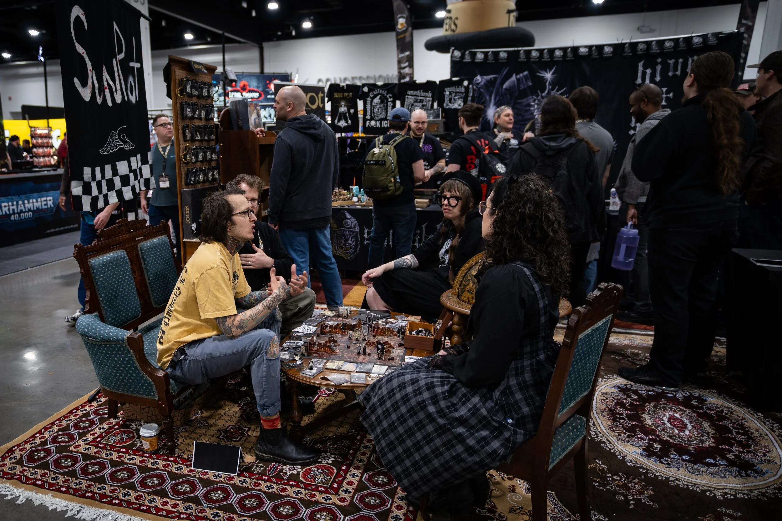 People gathered around a table playing a tabletop role-playing game at an indoor convention. Several chairs and a decorative rug are visible, along with game miniatures and cards on the table.