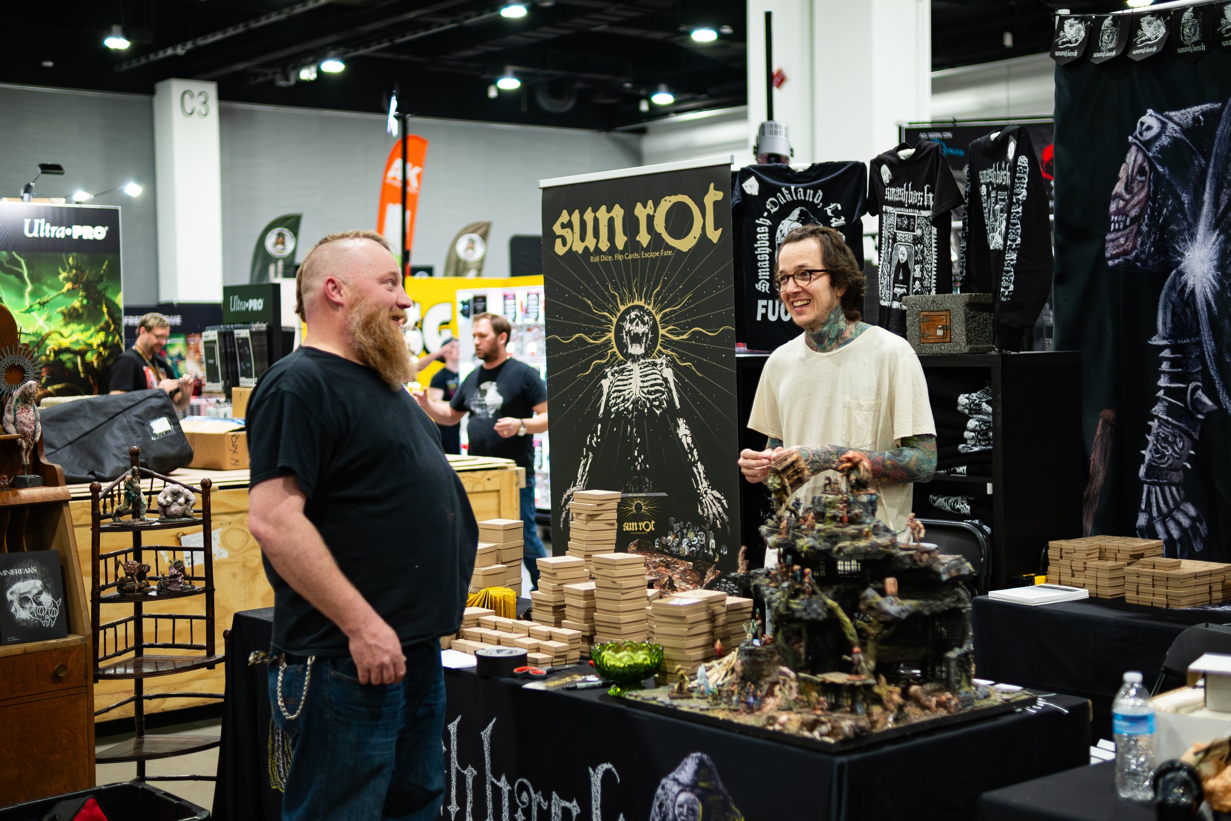 Two men are having a conversation at a booth during a convention, with various fantasy and gaming merchandise, including a prominent sign for 'Sunrot,' in the background.
