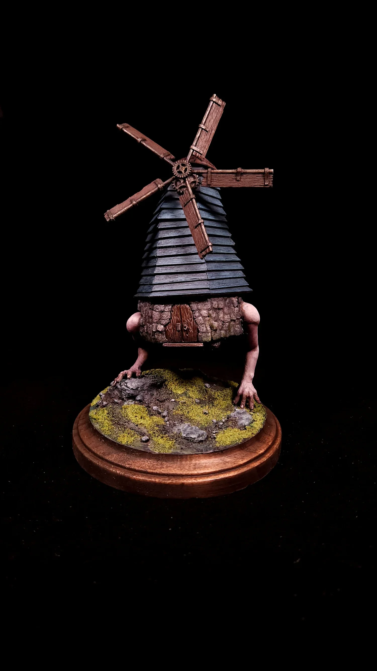 A whimsical sculpture of a windmill with human legs, standing on a round wooden base with moss and stones, against a black background.