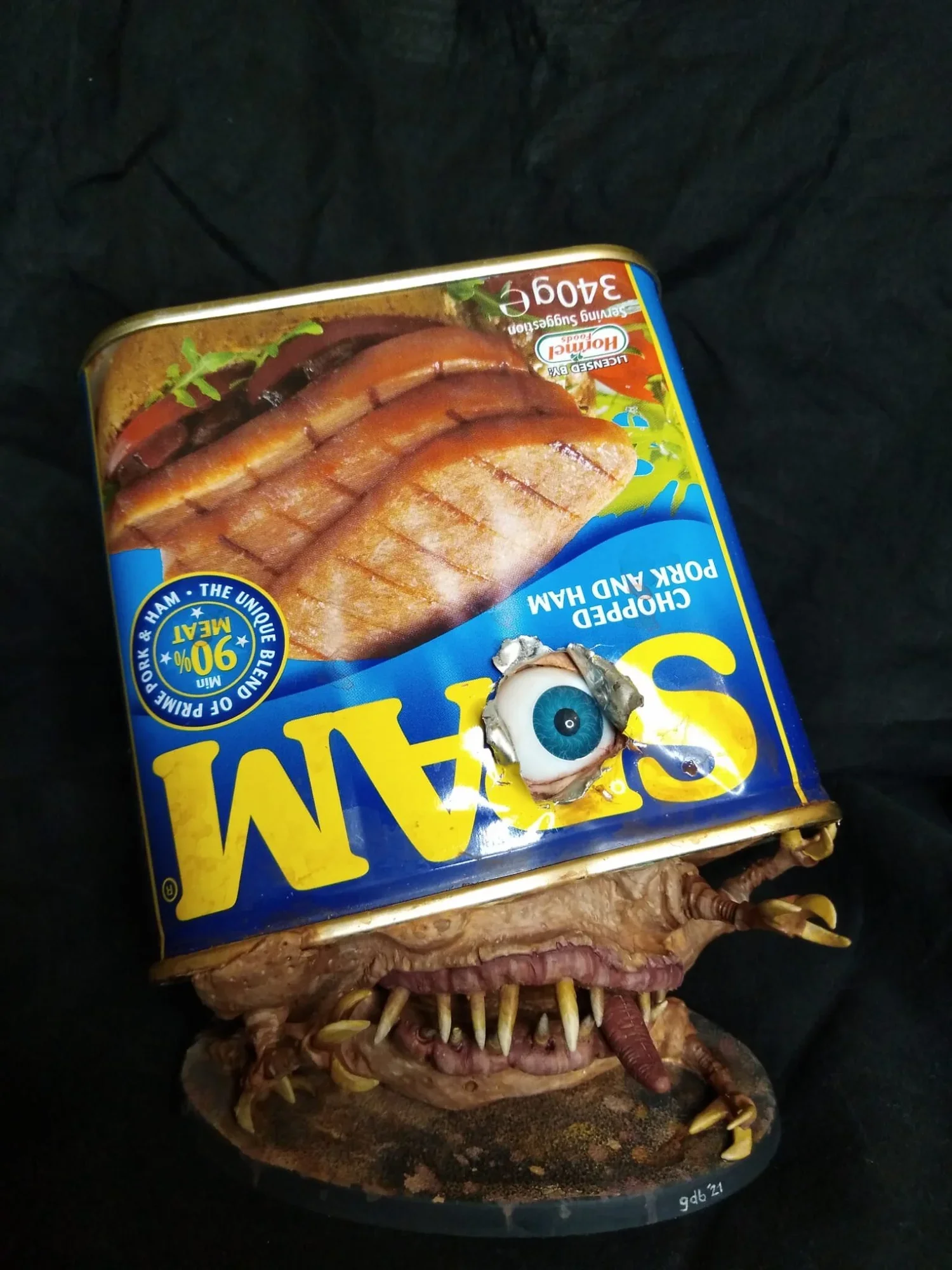 A surreal image combining a can of Spam with the head of a monster or creature, featuring sharp teeth, claws, and a single large eye on the can.