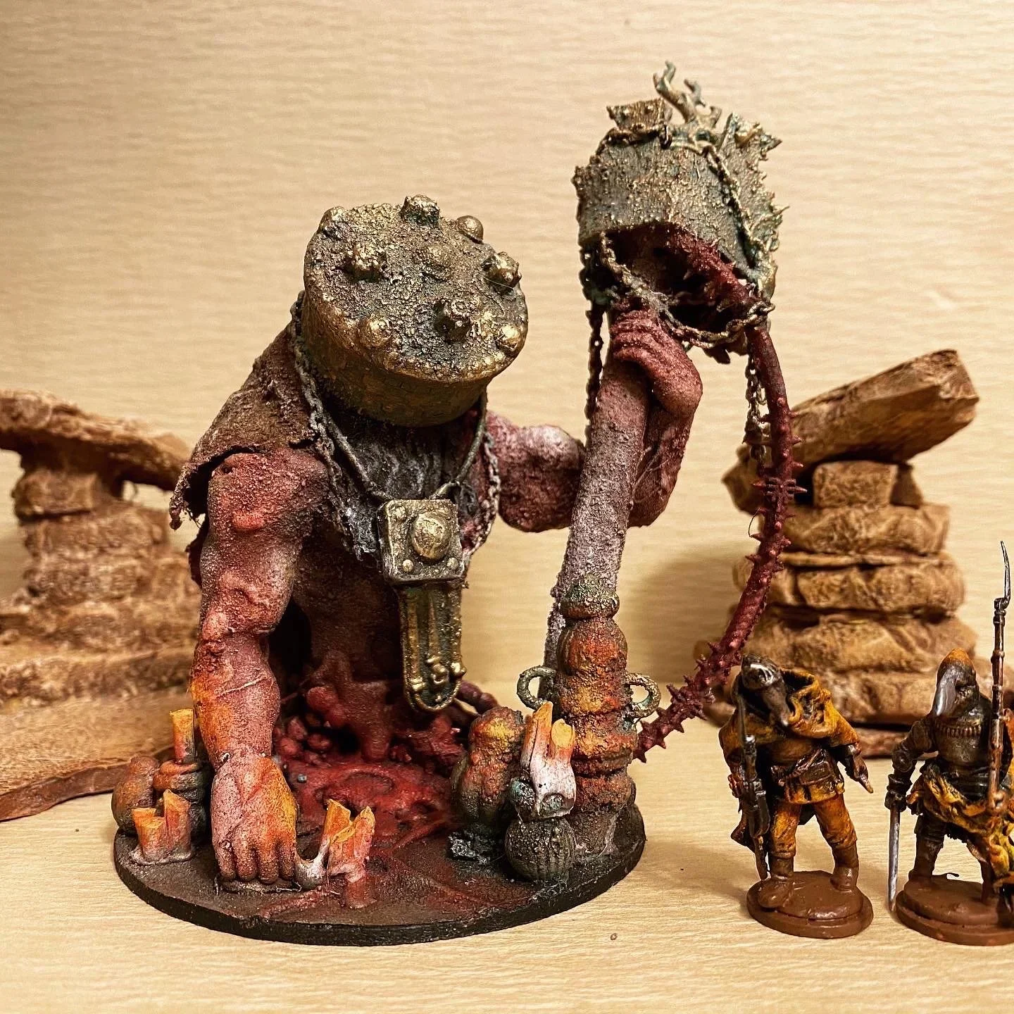 A collection of detailed miniature figurines, including a large figure with a textured head, reddish skin, and accessories, and two smaller soldiers in armor holding weapons, set against a neutral background with rocks.