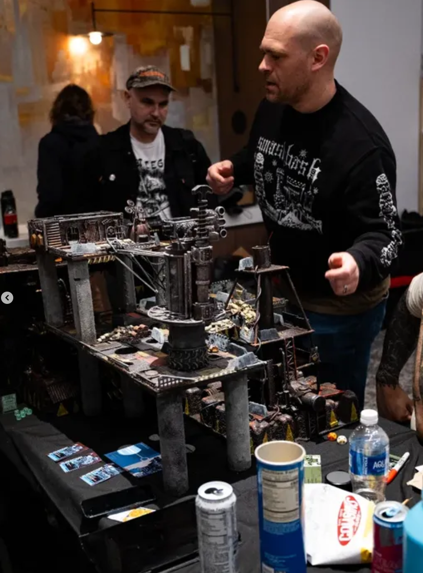 Three men in a room playing a tabletop miniature game with a detailed cityscape terrain. One man is actively engaged, while the others observe.