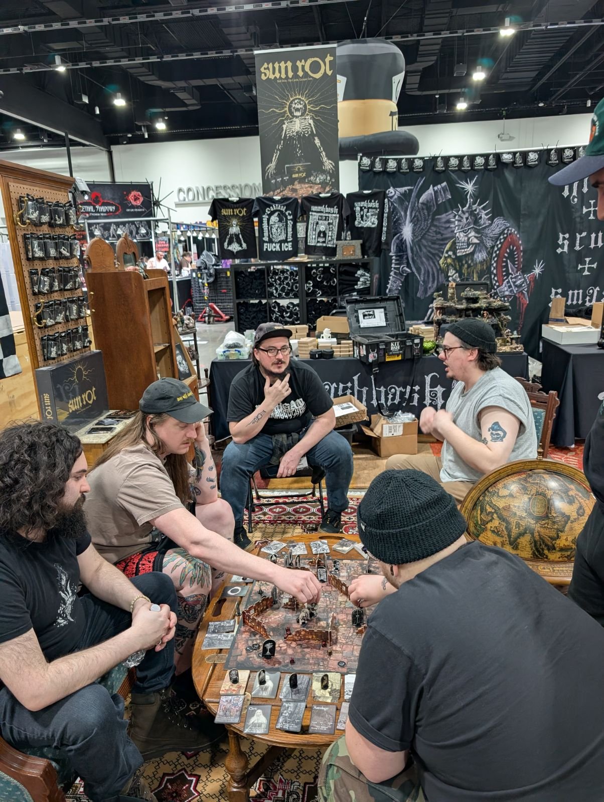 People playing a tabletop role-playing game at a convention booth with merchandise and artwork in the background.