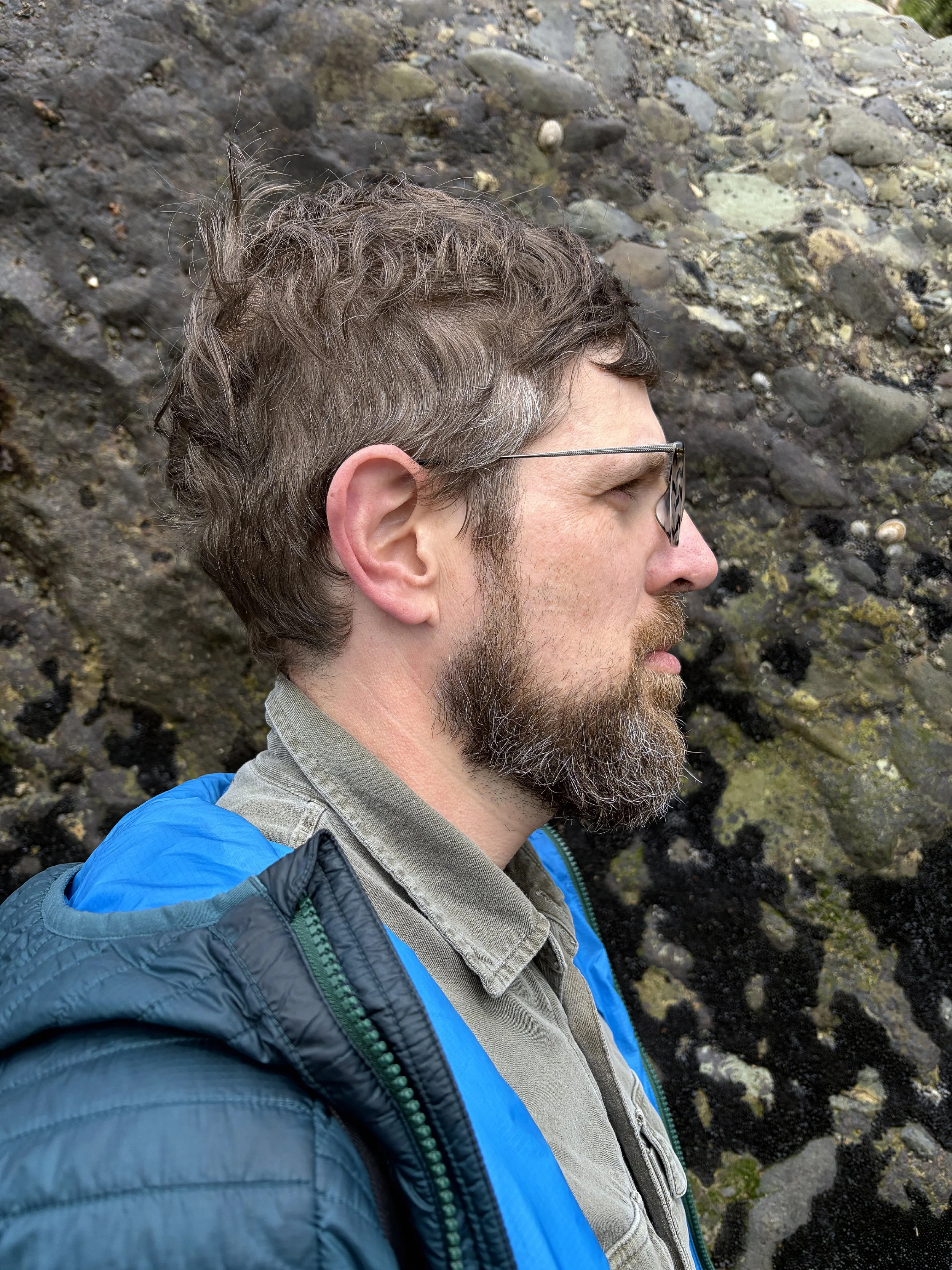 Side profile of a man with glasses, brown hair, and a beard, wearing a blue jacket and a gray shirt, standing in front of a rocky surface.