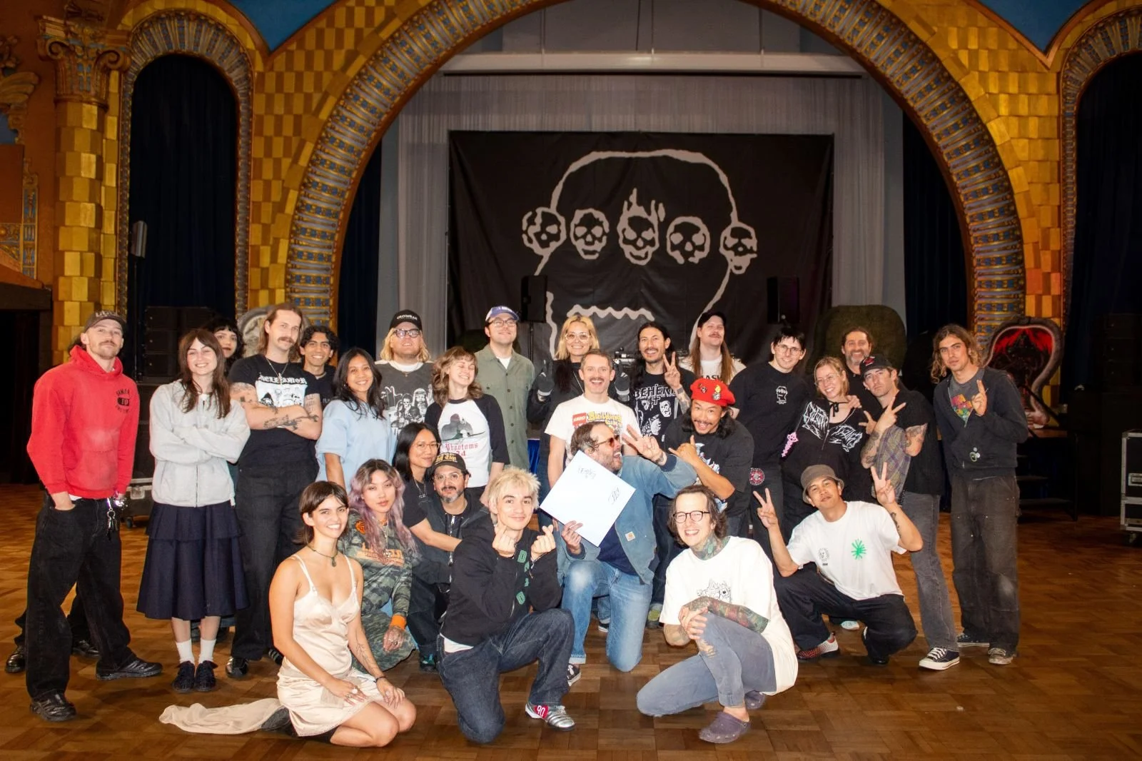 A large group of diverse people posing for a group photo in an indoor venue with a stage and a black backdrop featuring a white skull with multiple eyes. The group is smiling, making peace signs, and some are wearing casual or band-themed clothing.