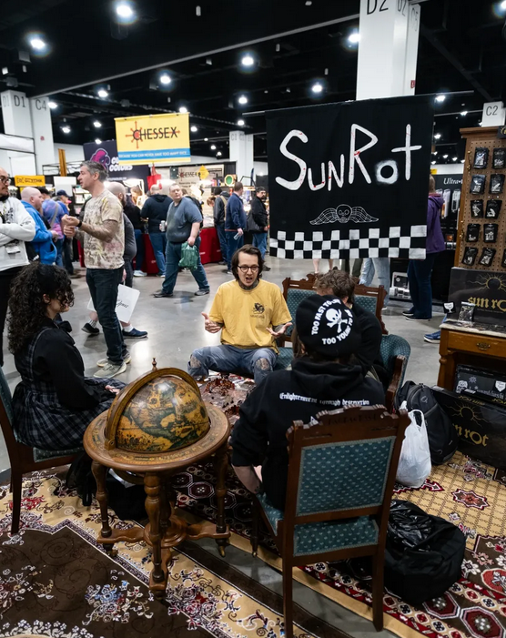 Four people sitting and talking at a convention or trade show booth with a large globe and vintage furniture, surrounded by other attendees and booths in the background.