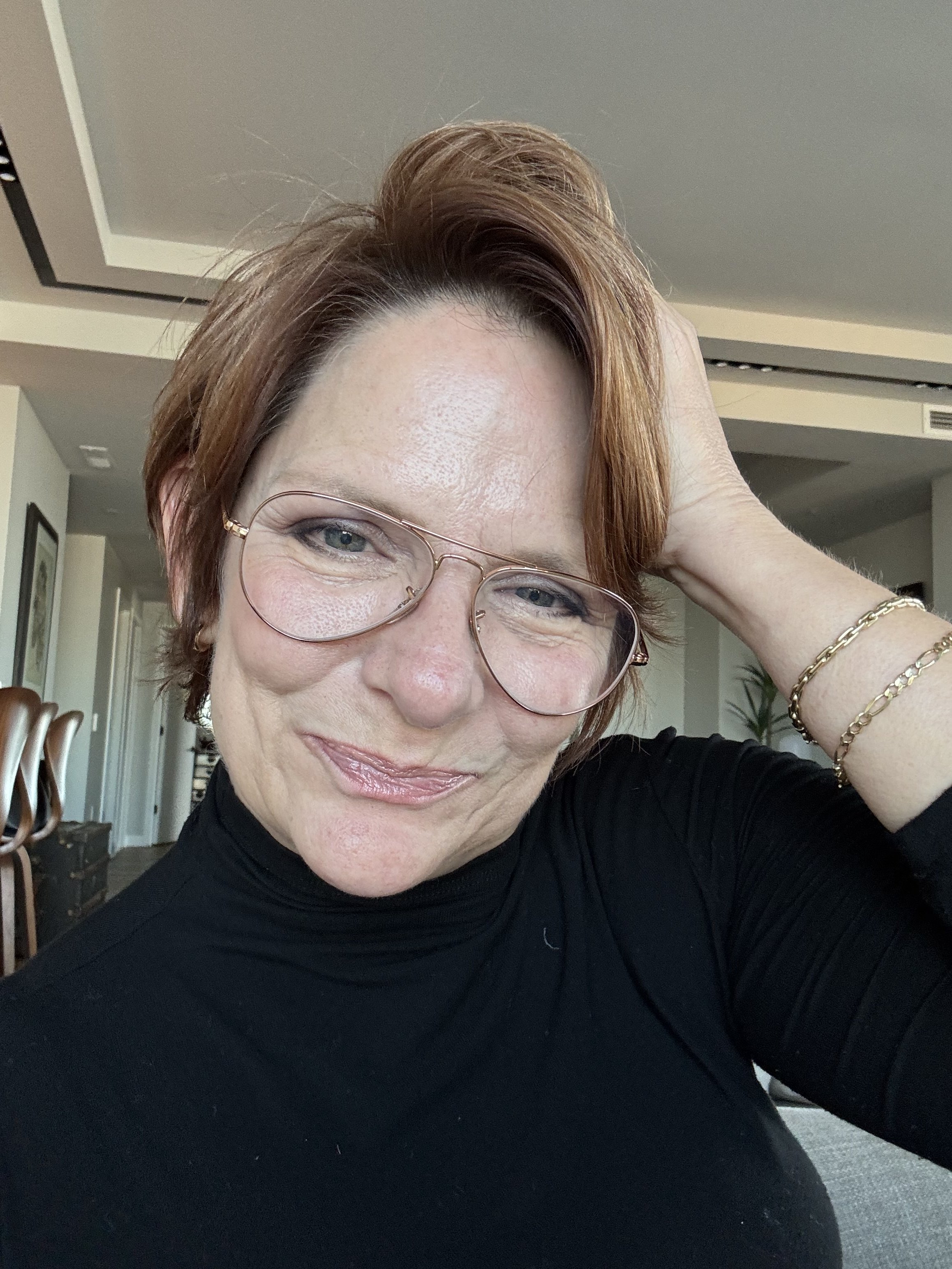 Close-up of a woman with short auburn hair and glasses, smiling and holding her hand behind her head, wearing a black turtleneck and jewelry, in a well-lit room with framed pictures and a plant in the background.