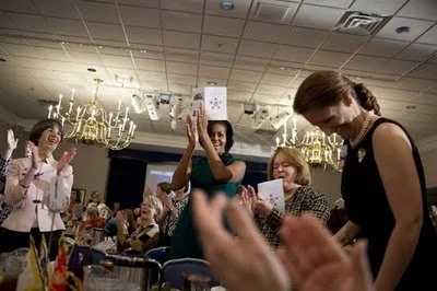 Women celebrating at an event, applauding and clapping, in a decorated room with chandeliers and tables.