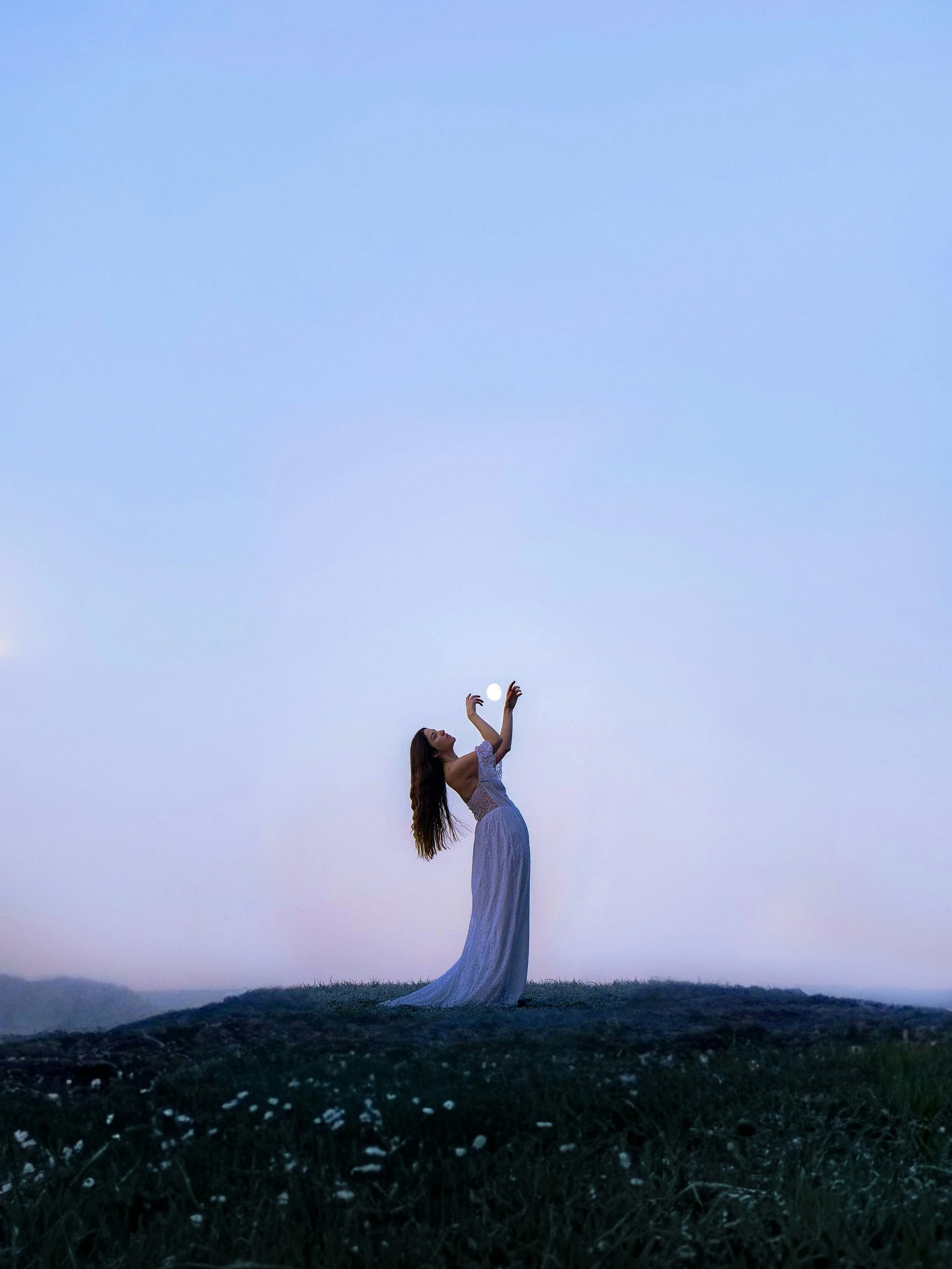A woman in a long dress stands on grass at dusk, reaching up toward the sky with her arms raised and fingers spread, as the sky darkens behind her.