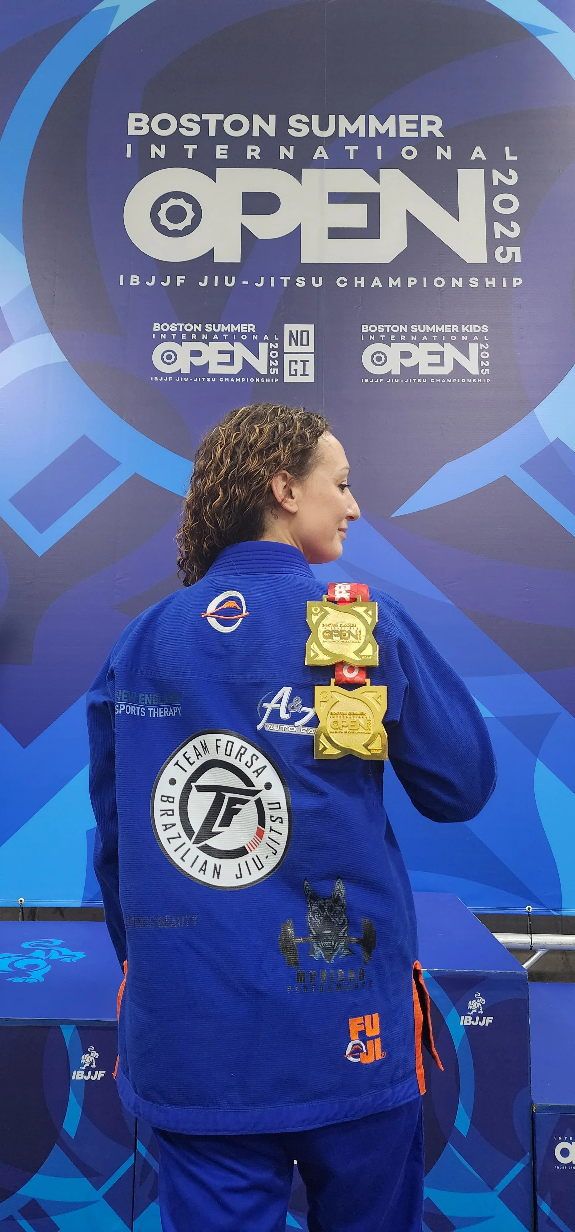 Dr. Jessica Camplese a Brazilian Jiu-Jitsu athlete wearing a blue gi with several patches standing in front of a blue and purple backdrop with event information for the 2025 Boston Summer International Open IBJJF Jiu-Jitsu Championship.
