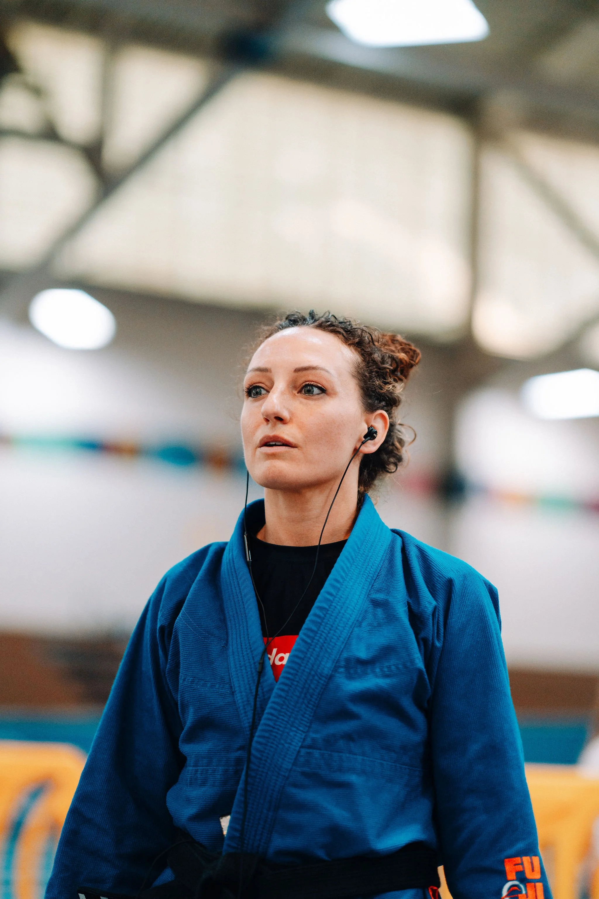 Dr. Jessica Camplese wearing a blue martial arts gi with patches, black belt, and earbud in her ear, practicing martial arts indoors.