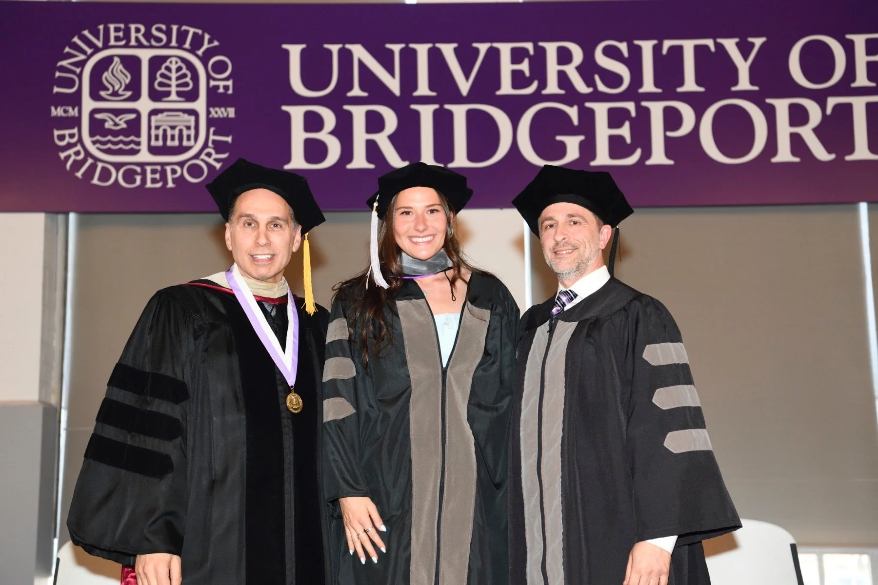 Three individuals in academic regalia standing in front of a banner that says 'University of Bridgeport' with the university's logo. They are smiling, and the middle person is a woman, flanked by two men.