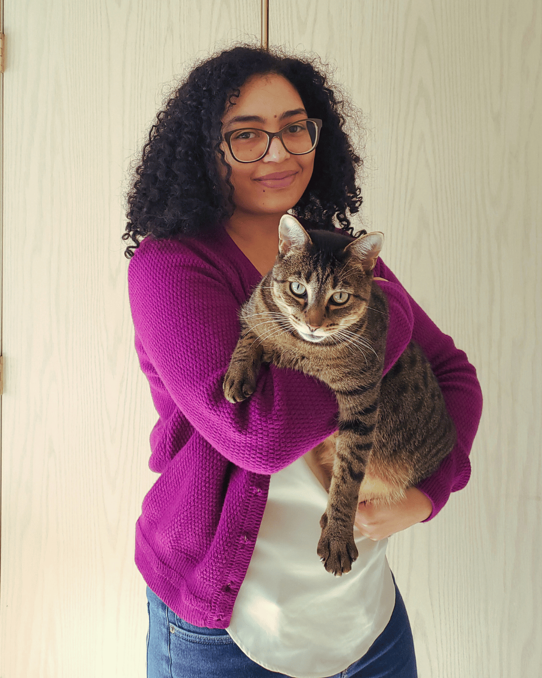 Isabelle smiling while slightly standing sideways, wearing a vibrant purple cardigan and holding her brown tabby cat, who is looking at the camera with one arm hanging loosely.