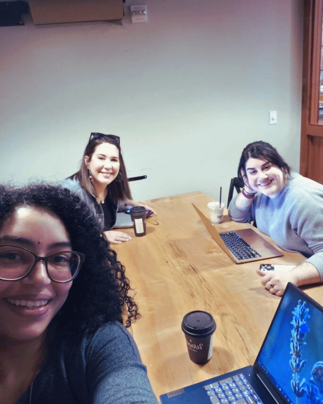 Selfie of three coworkers at a coworking table with laptops and coffee cups.