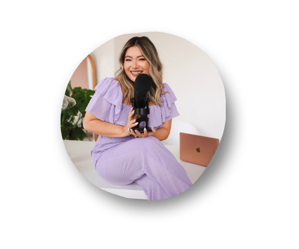 Smiling Ellen Yin, a Founder & podcast host of Cubicle to CEO, wearing a lilac outfit and sitting at a table with her legs crossed, holding a black Blue Yeti microphone. In the background, a laptop, plants, and a mirror are visible.