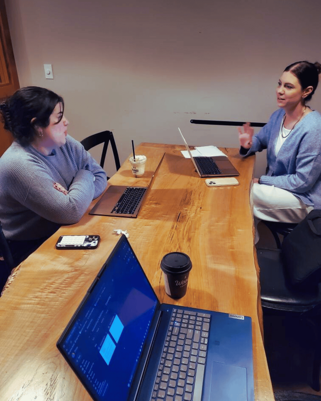 Two people having a conversation at a Monday Muse, with laptops and coffee cups on the table.