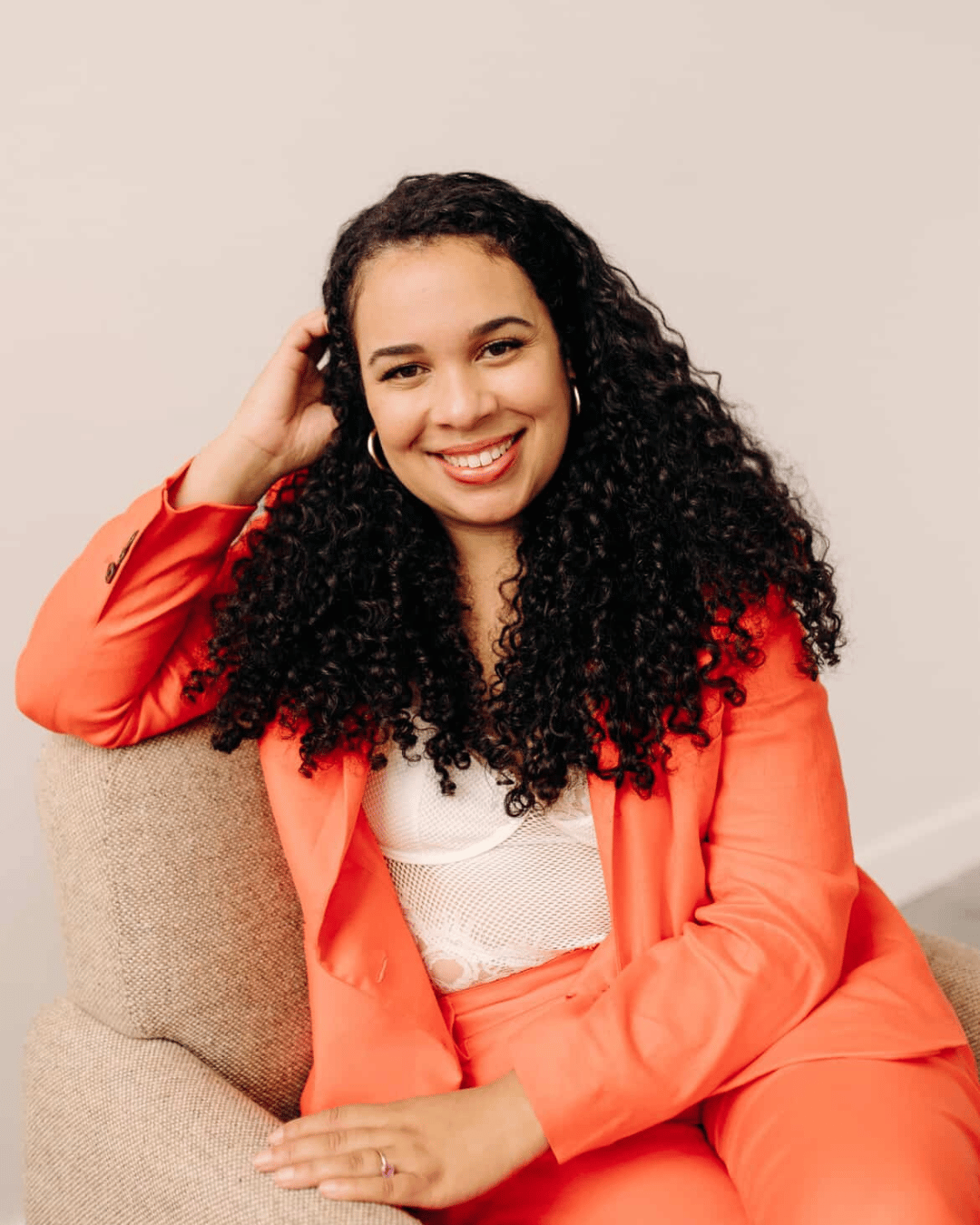 Image of Simone McNish, a DEI + Marketing Consultant wearing an orange suit, while sitting on a cream chair and resting her right elbow on the top of the chair.