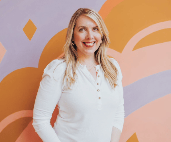 A smiling woman with blonde hair, wearing a white shirt, standing in front of a colorful abstract background.
