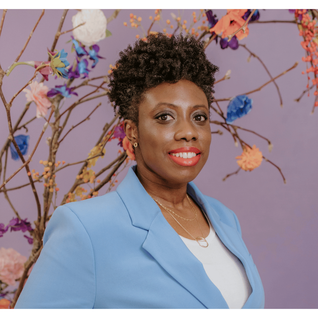 A woman with short, curly black hair, wearing a light blue blazer and a white top, standing in front of a purple background decorated with colorful flowers and branches.