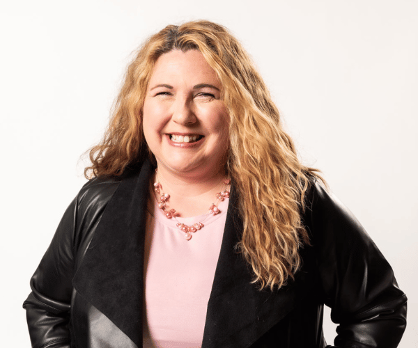 A woman with curly blonde hair smiling and wearing a pink top, a black leather jacket, and a pink beaded necklace.