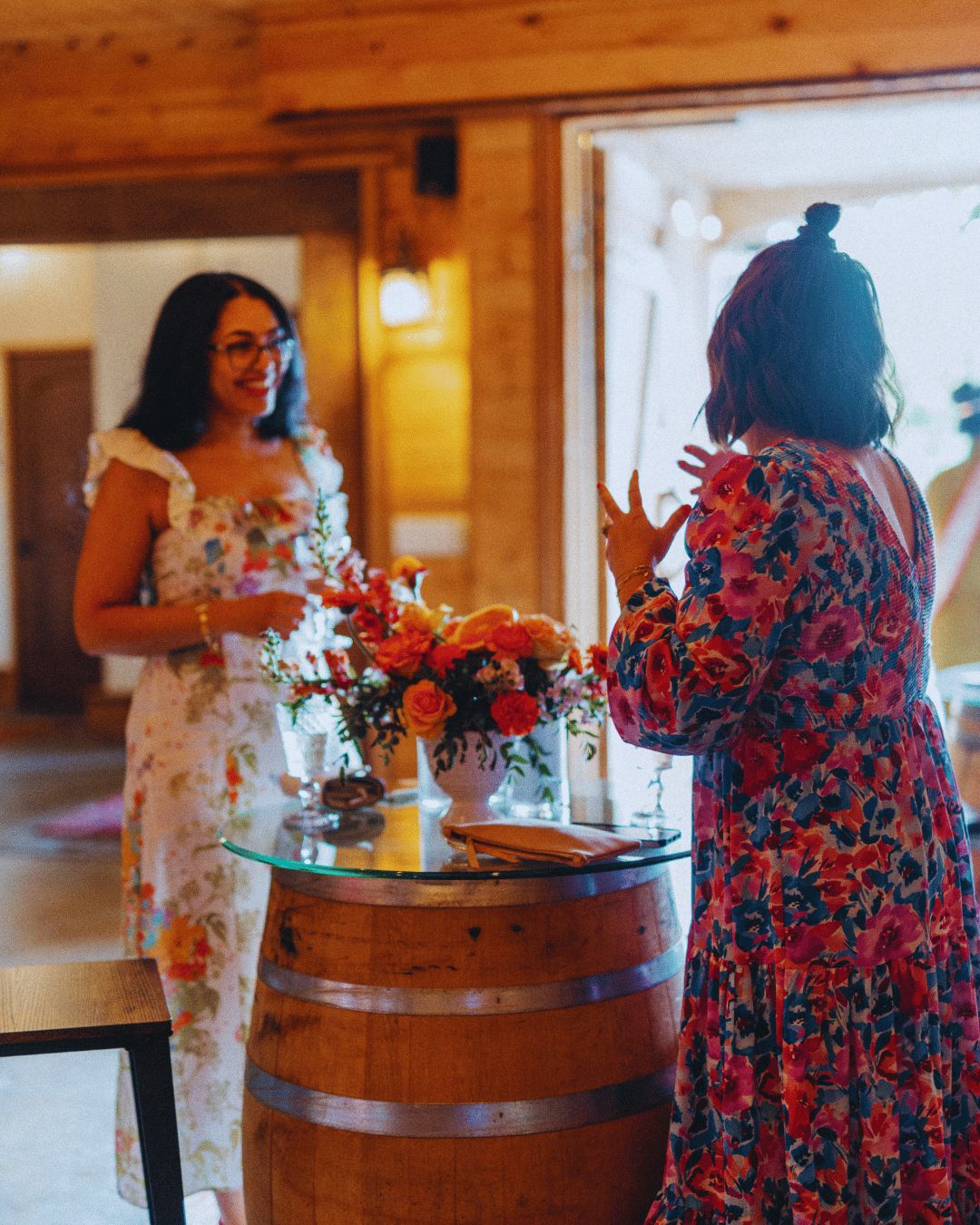 Isabelle smiling and engaged in a deep conversation with someone, their back to the camera, standing around a small tall circle table with vibrant orange flowers at a business event.