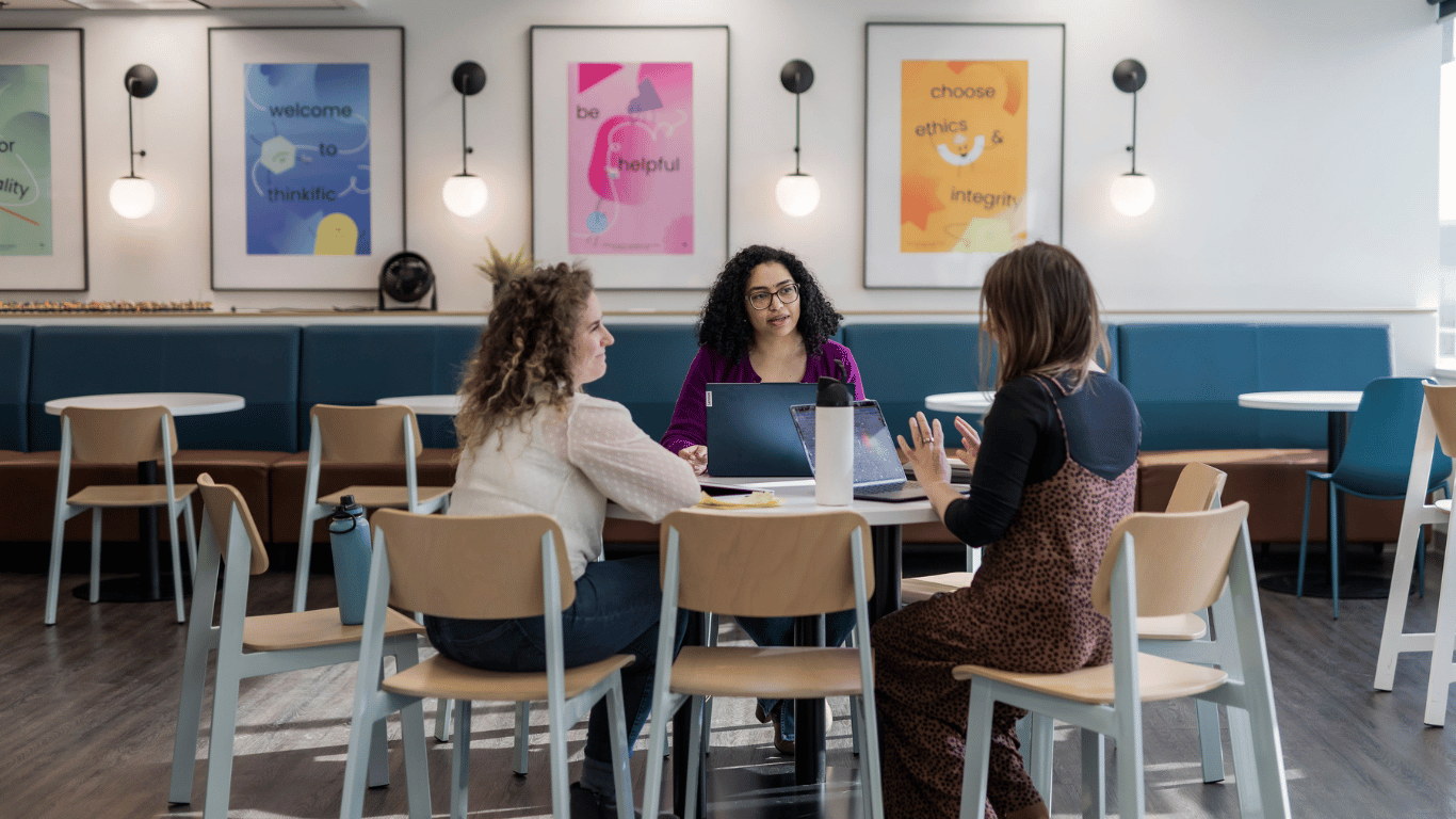 Isabelle sitting at a round table with two business owners, engaging in a conversation while working on their laptops and taking notes in notebooks. Collaboration and strategy session for business growth.