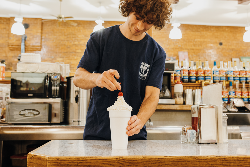 A man makes a milkshake behind a counter, topping it with a cherry