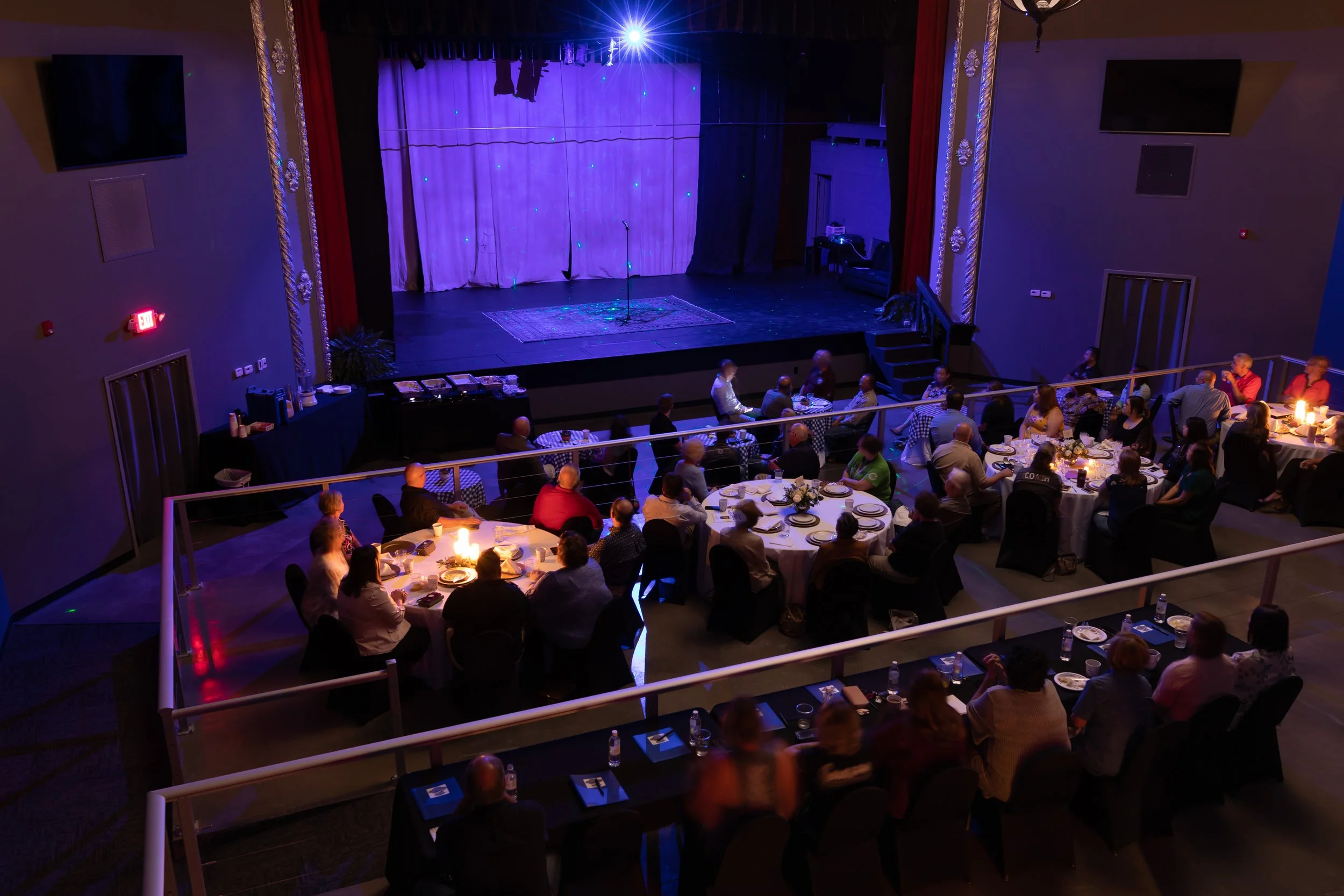 People sit at round tables in front of a stage lit blue. Everyone has a plate in front of them.