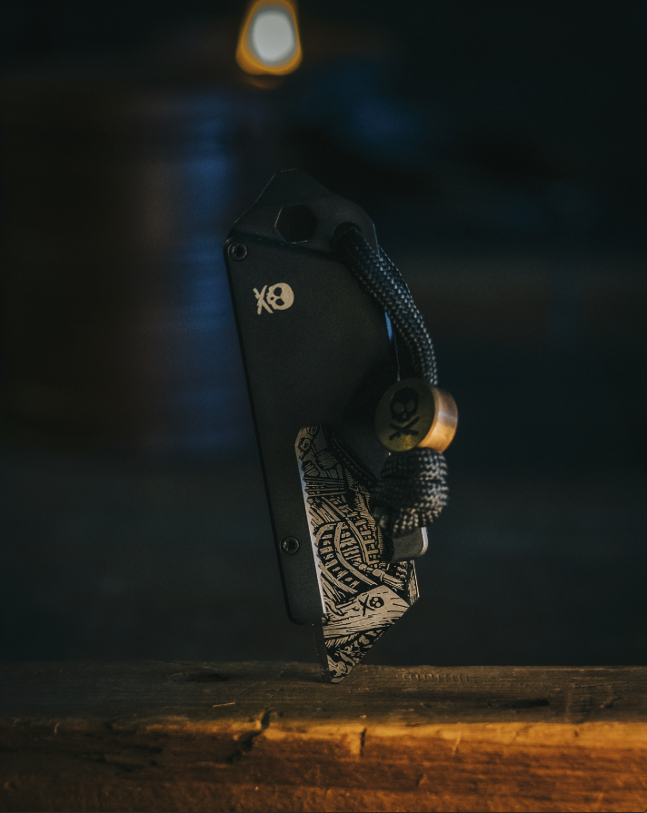 Close-up of a black pocket knife with a skull logo and a silver engraved pattern on the blade, attached to a black and gold bead lanyard resting on a wooden surface.