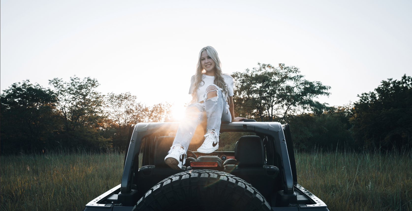 A young woman with long blonde hair, wearing a white distressed t-shirt and ripped jeans, sitting on the top of a black Jeep with a spare tire, in an open grassy field during sunset, smiling at the camera.