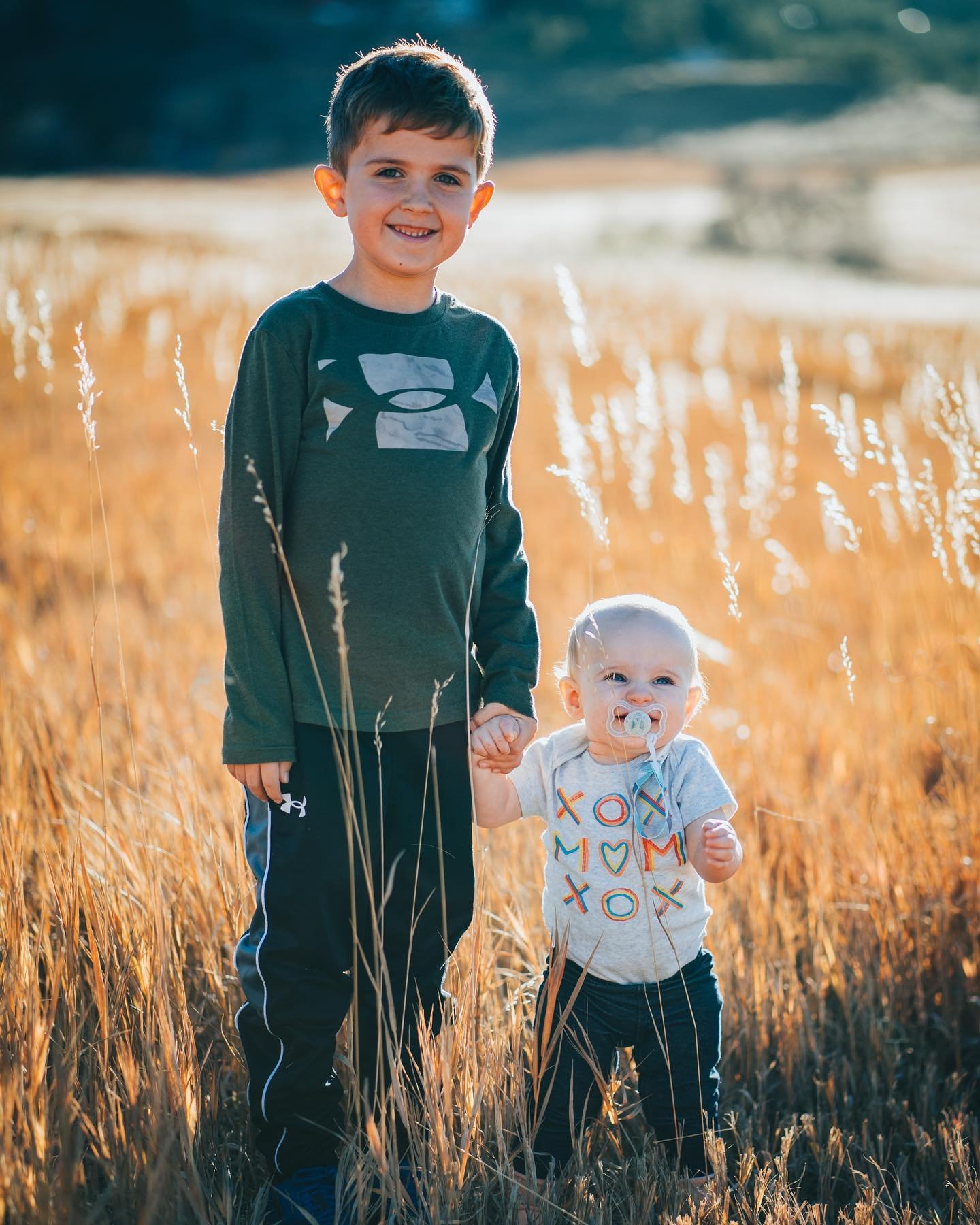 A young boy and a toddler girl holding hands in a golden grassy field during sunset, with the girl having a pacifier, and both children smiling.