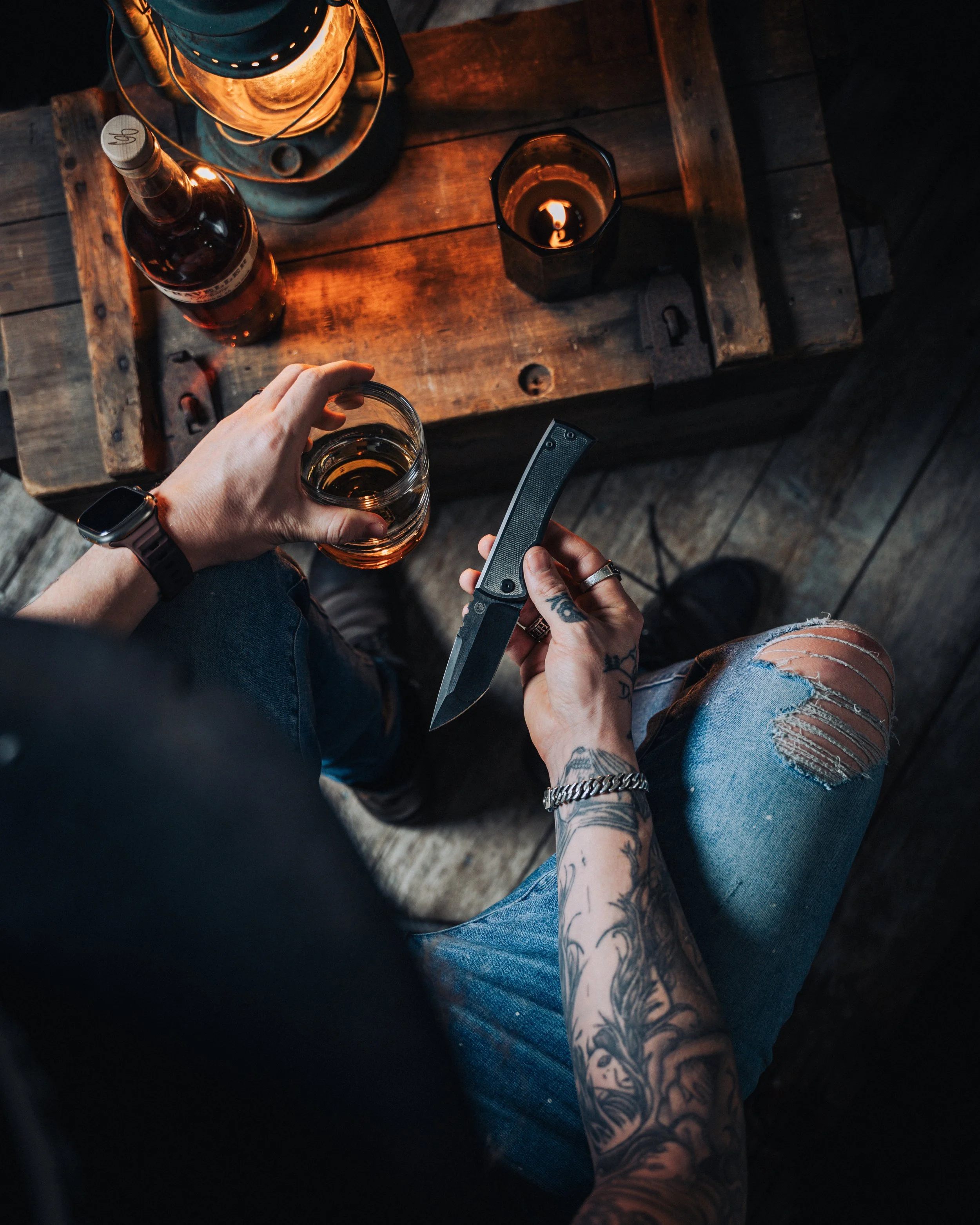 Person with tattoos holding a folding knife in one hand and a glass of amber-colored beverage in the other, sitting at a rustic wooden table with a lantern, a bottle, and a candle.