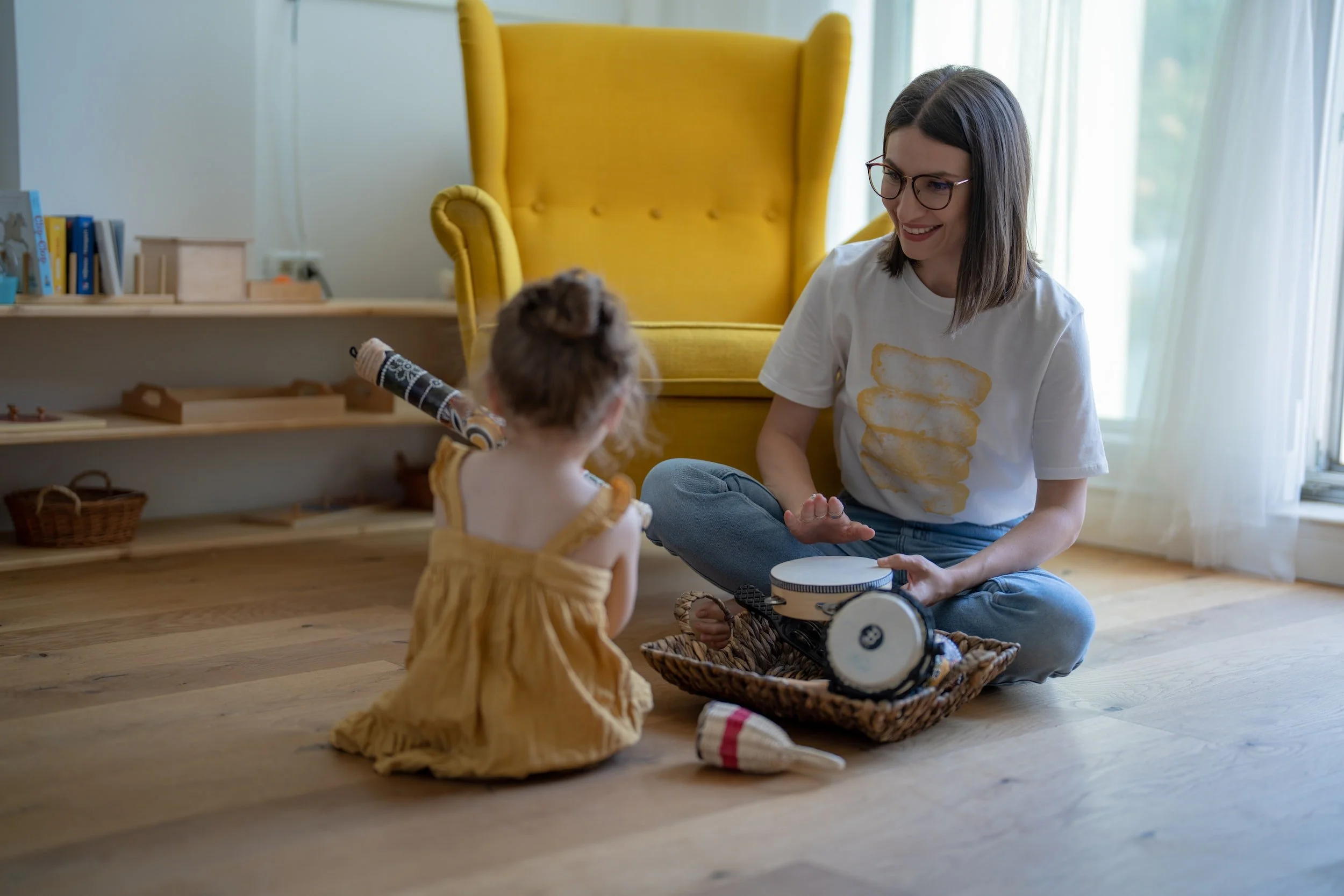 A woman and a young child sit on a wooden floor playing with musical instruments. The woman is holding a tambourine, and the child is holding a rainstick. A bright yellow armchair is in the background.