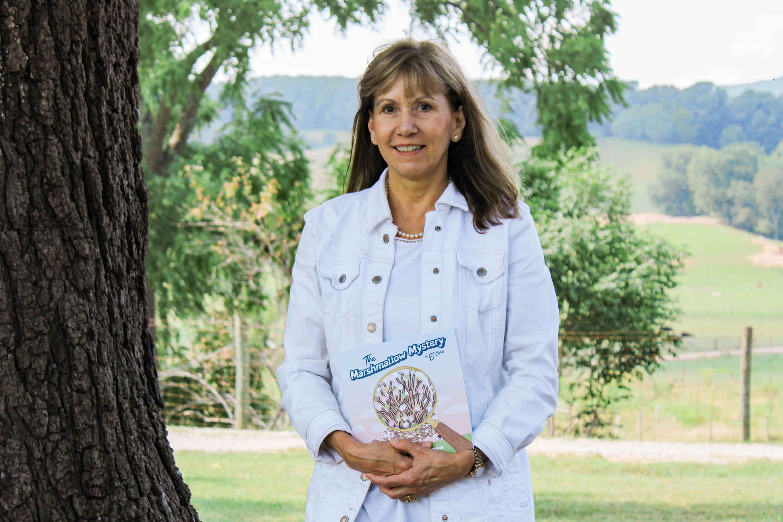 A woman standing outdoors near a tree, holding a book titled 'The Marshmallow Mystery'. She is smiling, wearing a white jacket and in the background, there are trees and a scenic landscape with green hills.