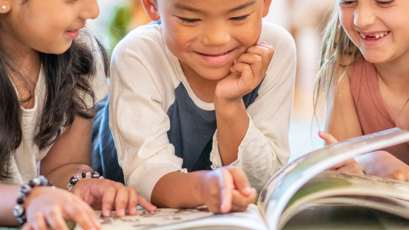 Three children reading a book together, smiling and looking engaged.