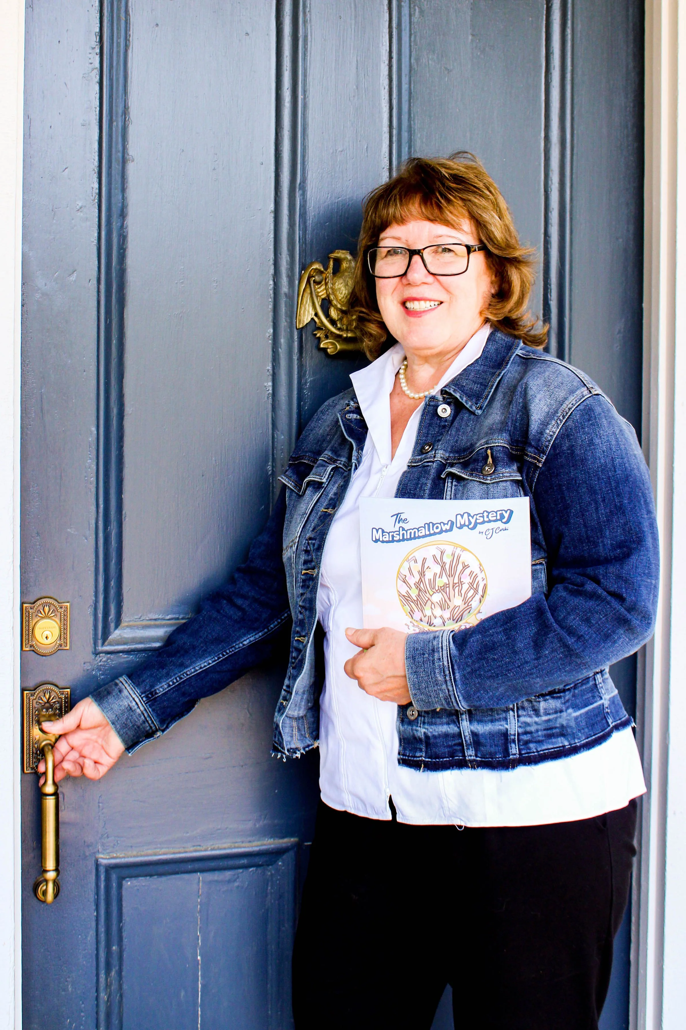 A woman with glasses and a pearl necklace, holding a book titled 'The Marshmallow Mystery,' standing at a blue front door.