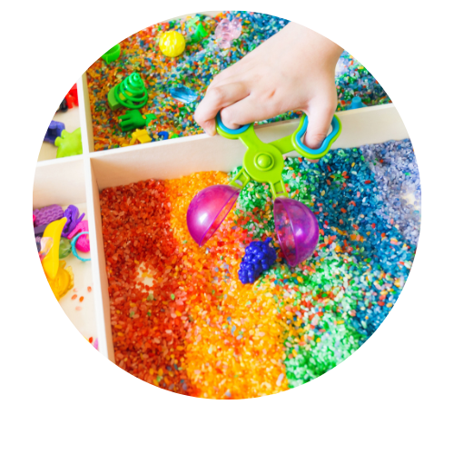 Hand holding a colorful claw tool above a divided sensory bin filled with rainbow-colored rice and toy shapes.